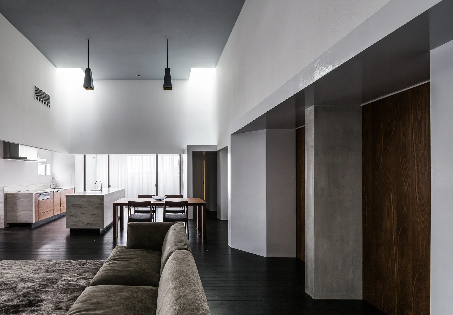 A minimalist open-plan living room and kitchen featuring a dark grey ceiling, dark wood floors, and recessed skylights above white walls.