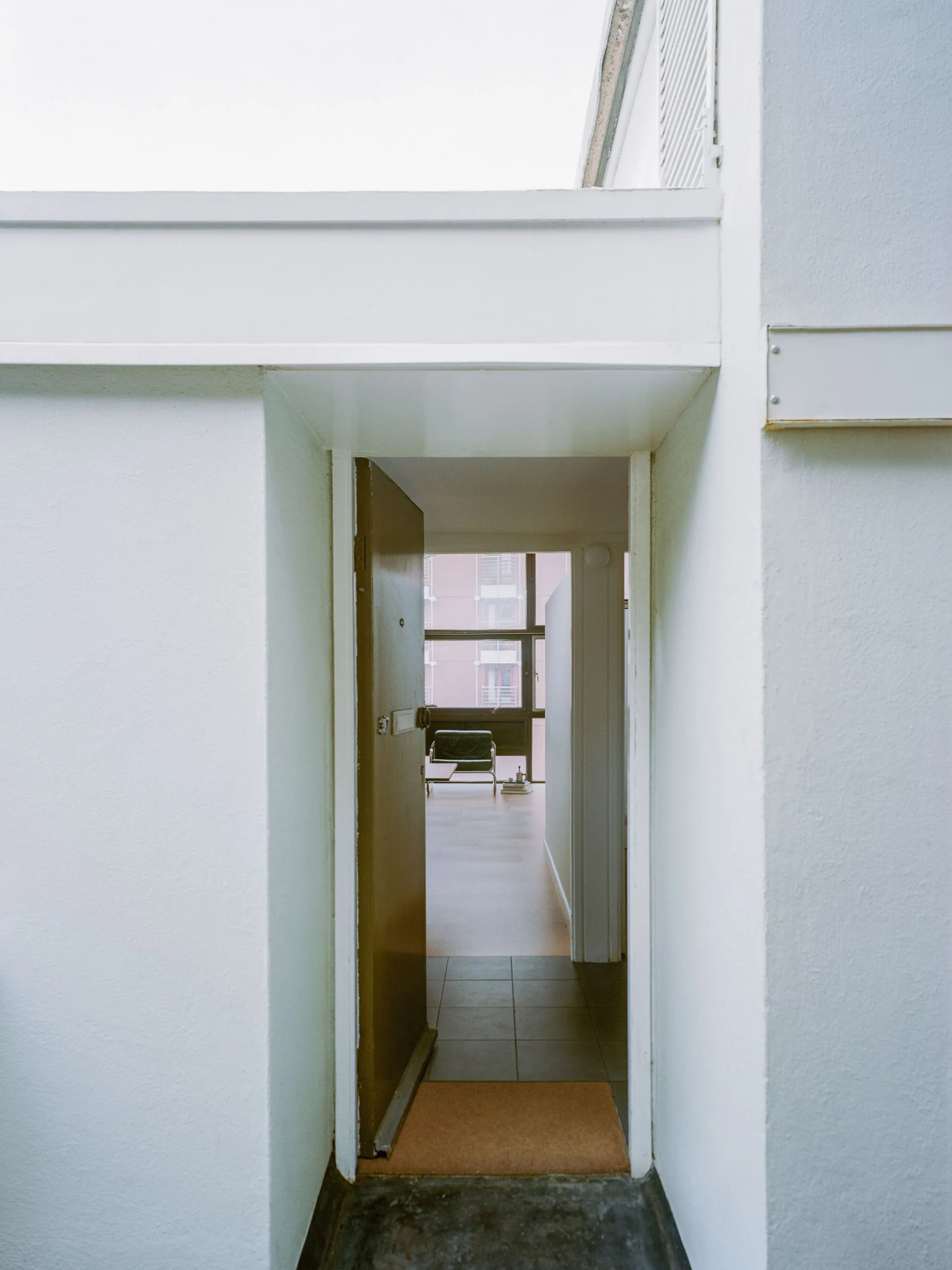 A view through an open tan door into a minimalist interior featuring white walls, gray floor tiles, and a sunlit living room with a large window.