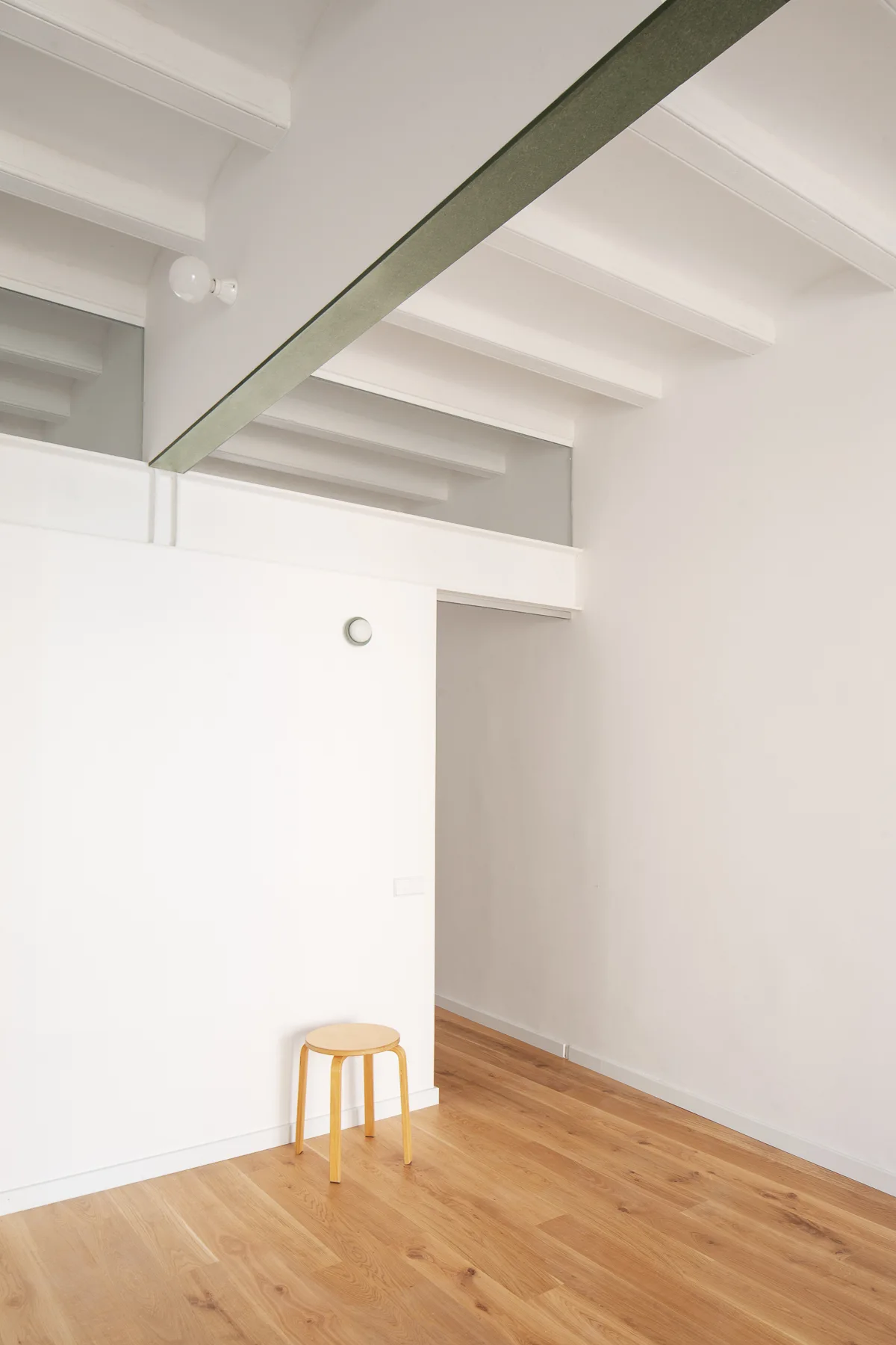 Minimalist white room featuring light wood flooring, a single wooden stool, an exposed dark ceiling beam, and a glass transom partition.