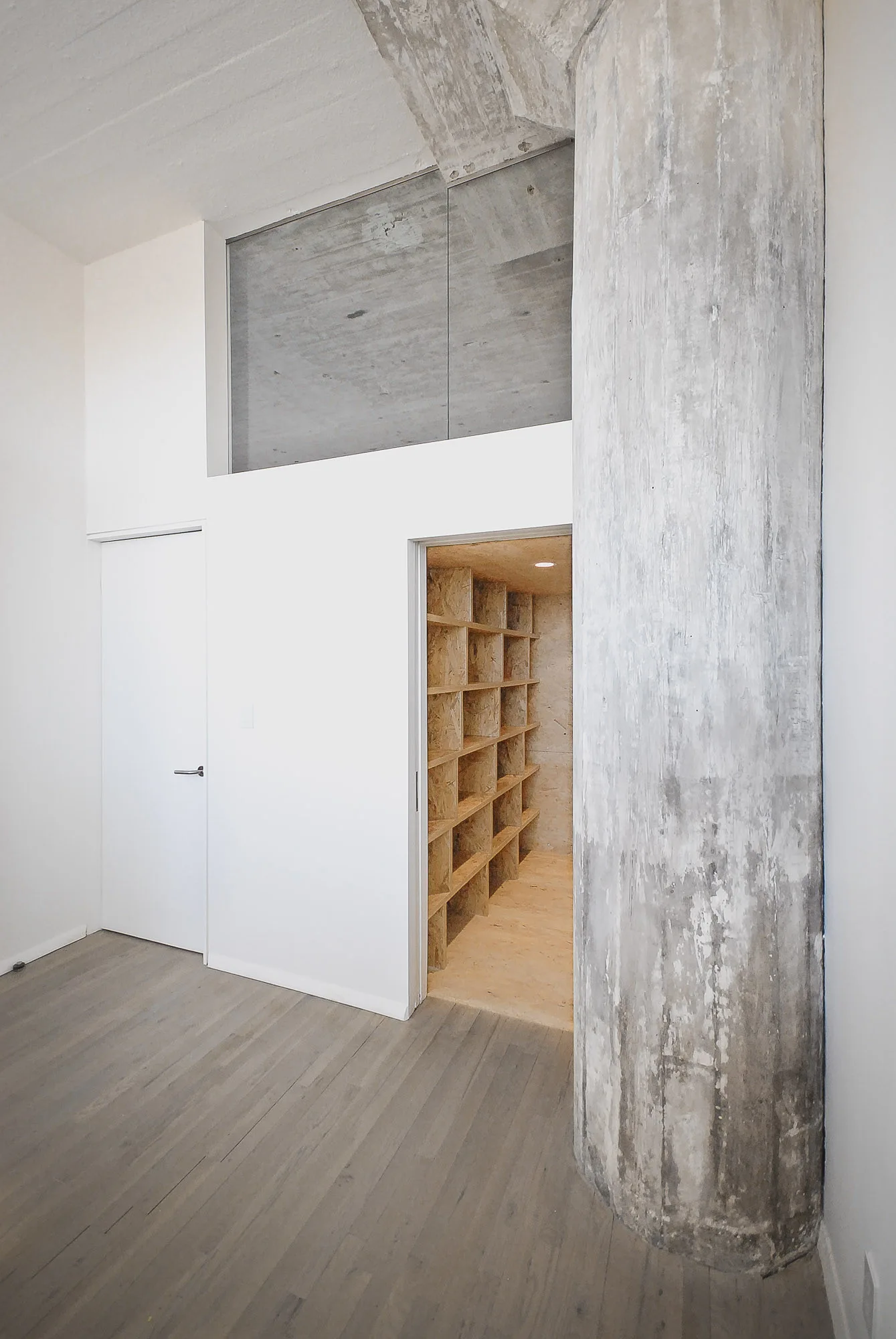 A minimalist room with a massive raw concrete column, gray wood floors, white walls, and an open doorway leading into an OSB-shelved walk-in closet.