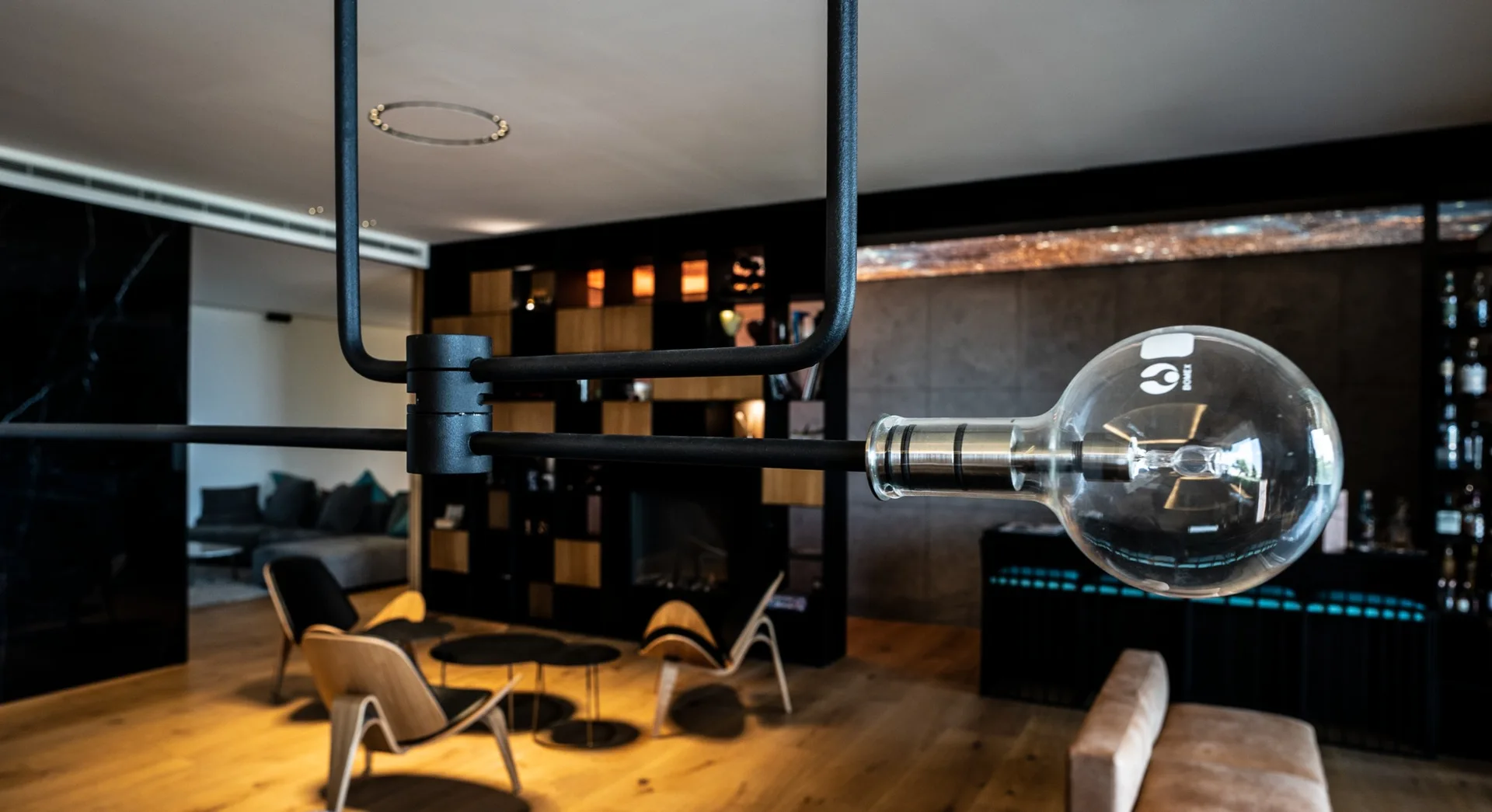 A close-up of a black metal pendant light with a clear bulb hangs in a modern living room featuring wood lounge chairs and dark built-in shelves.