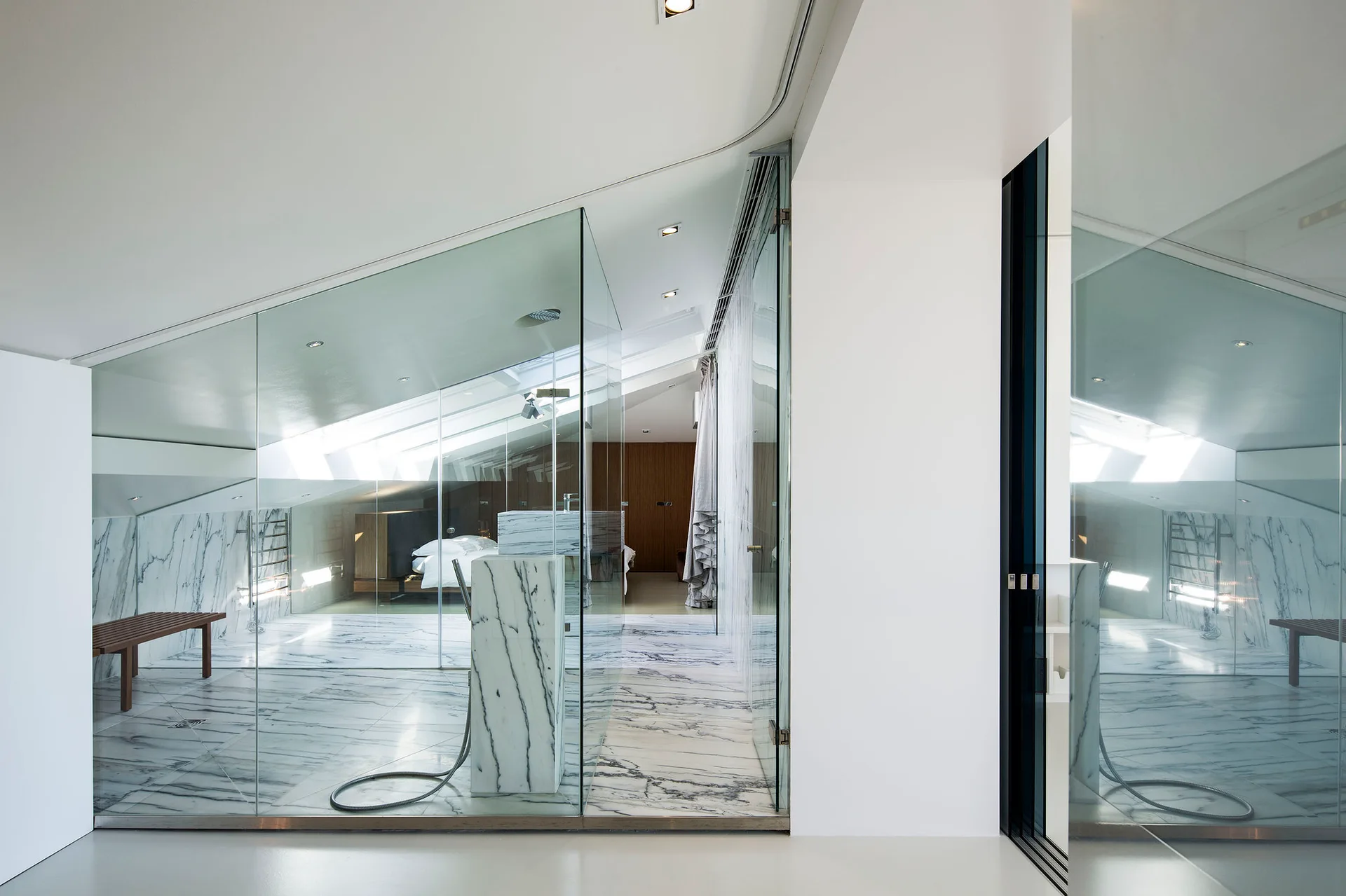 A modern master bathroom featuring white veined marble on the floor and walls with a frameless glass wall next to a bedroom under a sloped ceiling.