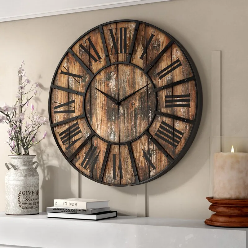 A large round distressed wood wall clock hangs on a beige wall above a white mantel styled with a milk jug vase, books, and a pillar candle.