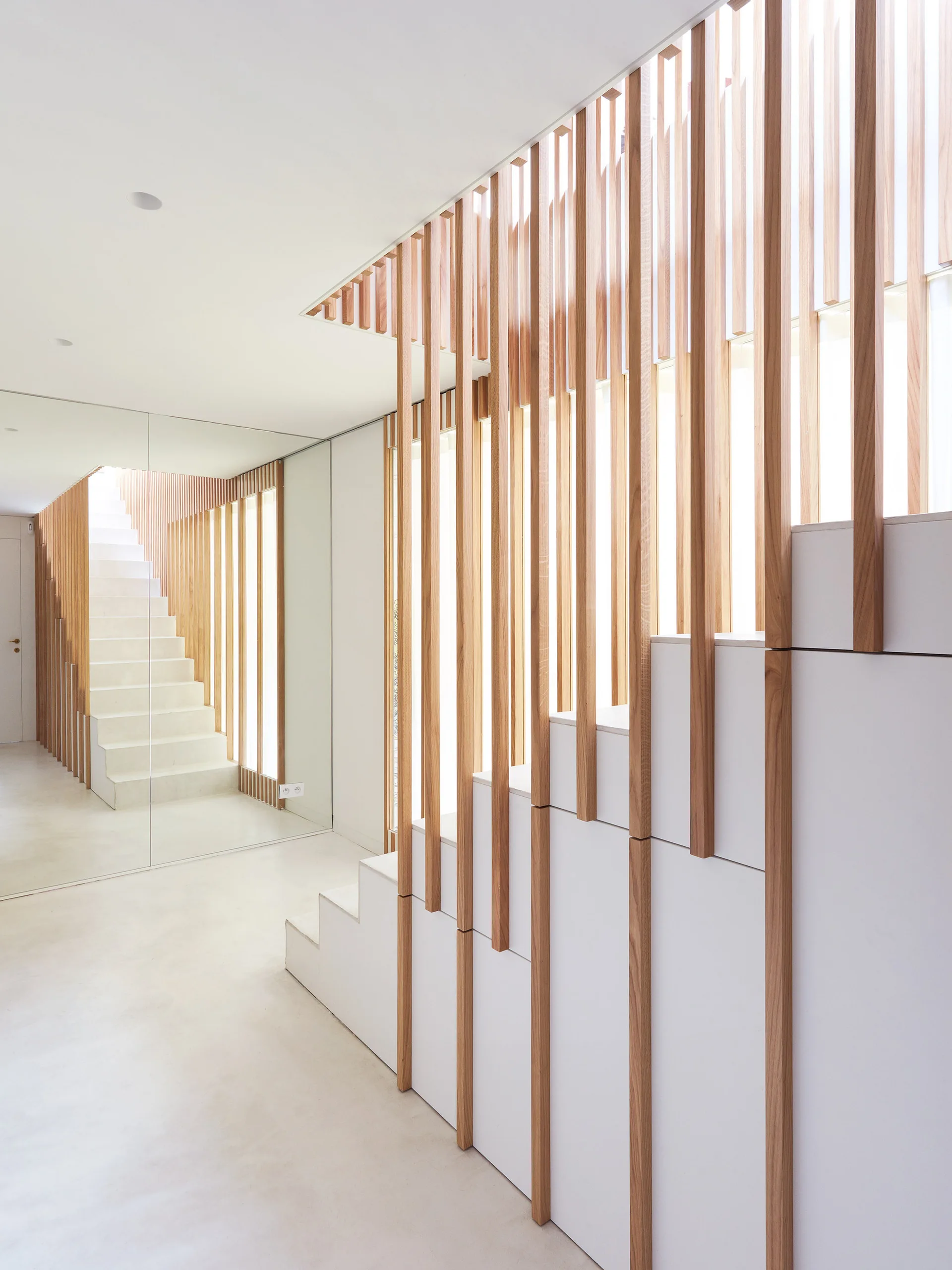 A white minimalist staircase features vertical light wood slats acting as a screen alongside a full-height mirrored wall.