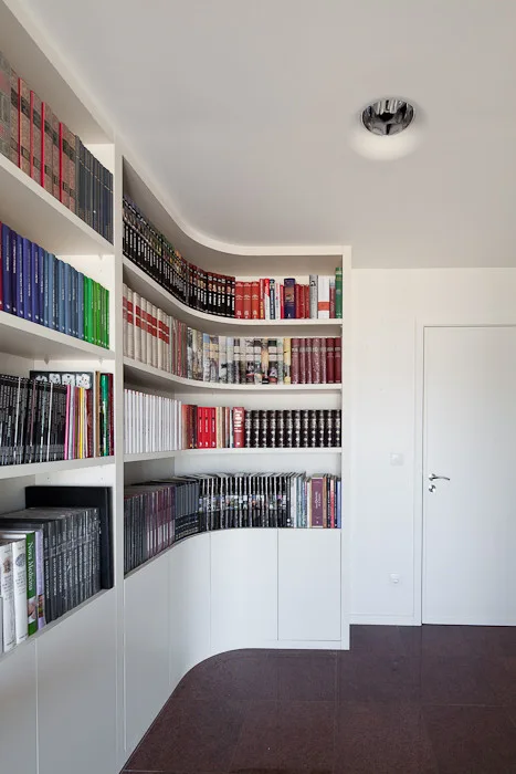 A custom curved white bookshelf filled with books wraps around a corner next to a white door in a modern room with dark tile floors.