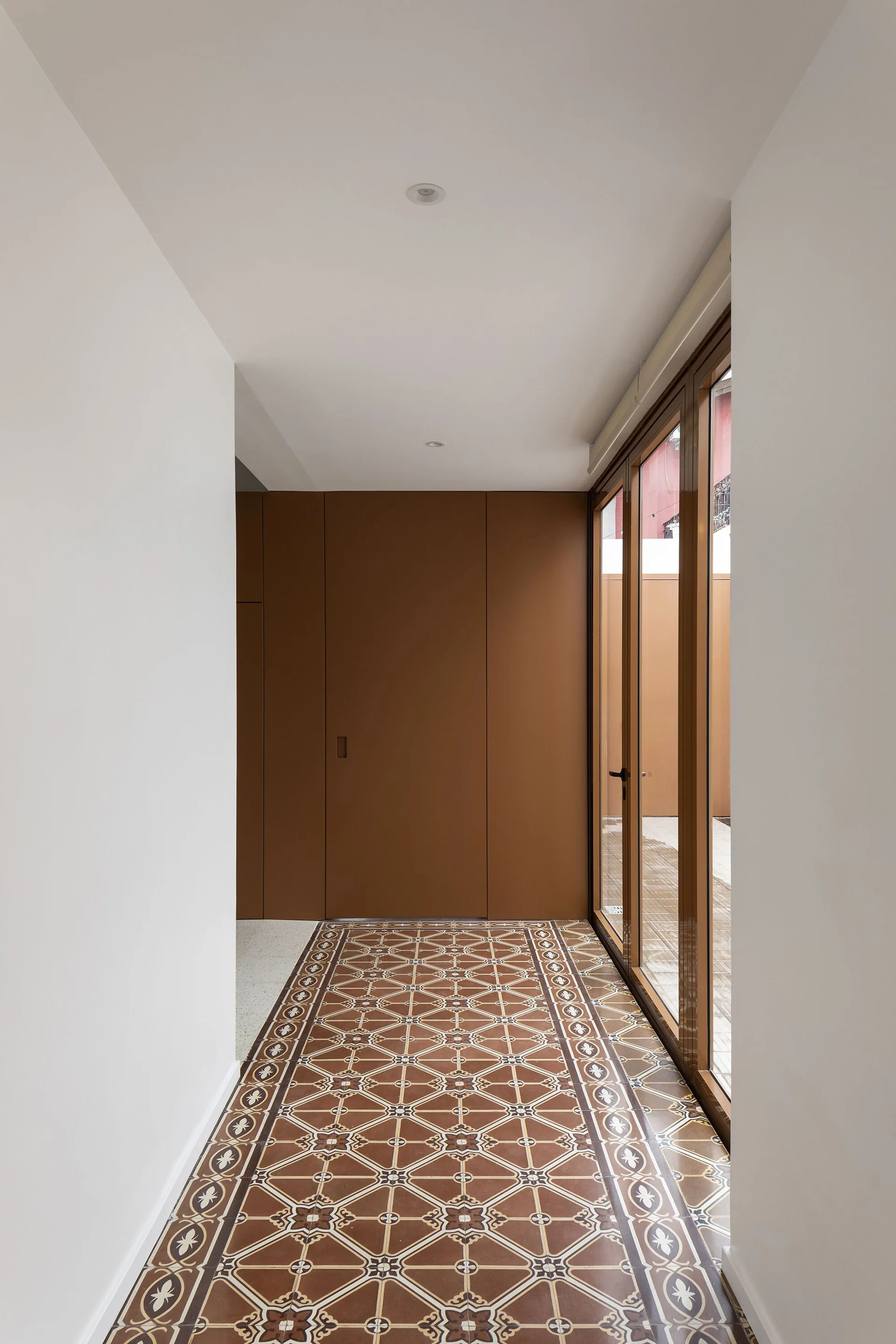 An interior hallway features brown geometric floor tiles, white walls, and a matching brown paneled wall alongside glass panel doors.
