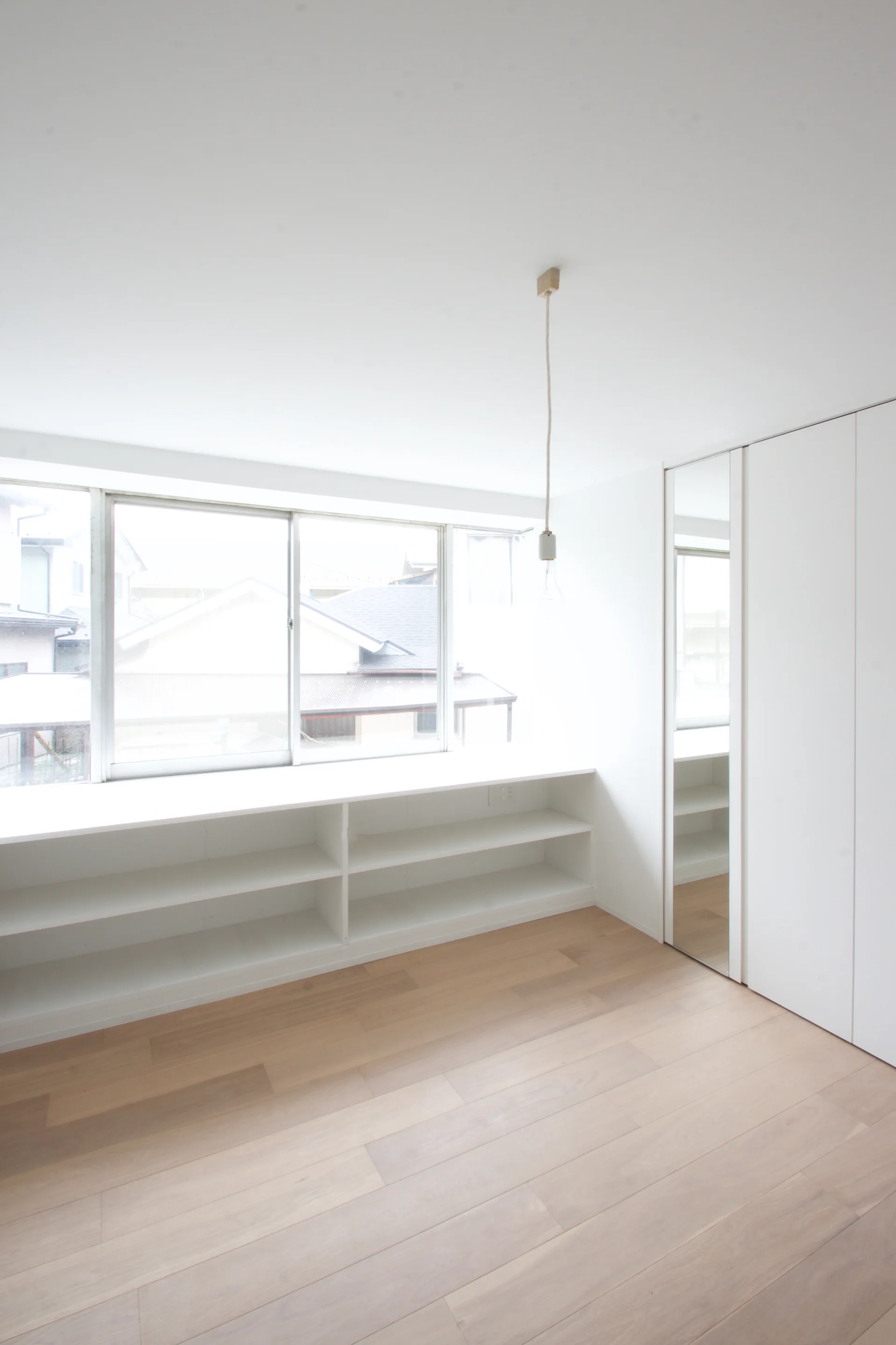 A minimalist white room featuring light wood flooring, a low built-in shelving unit under a wide window, and a tall wardrobe with a mirrored door.