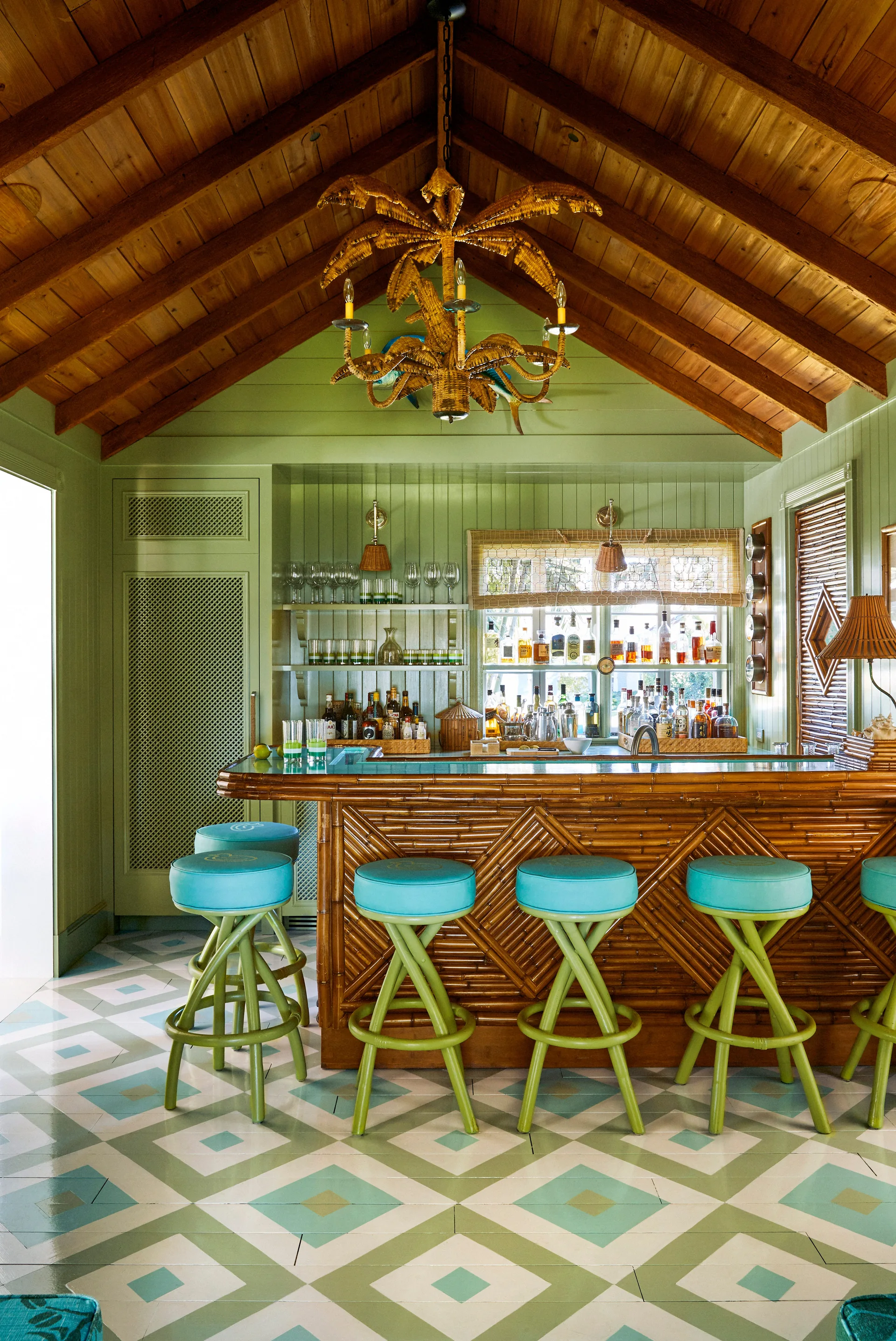 A tropical home bar features a bamboo counter with bright turquoise stools, sage green wood walls, a painted diamond floor, and a vaulted wood ceiling.