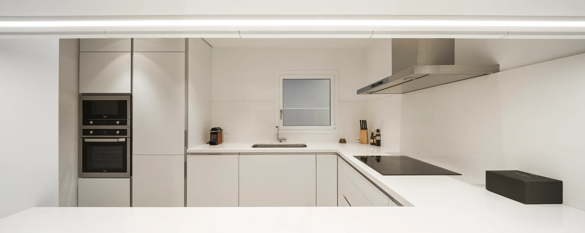A minimalist white kitchen features handleless cabinets, a built-in double oven, and a stainless steel range hood above a black cooktop.