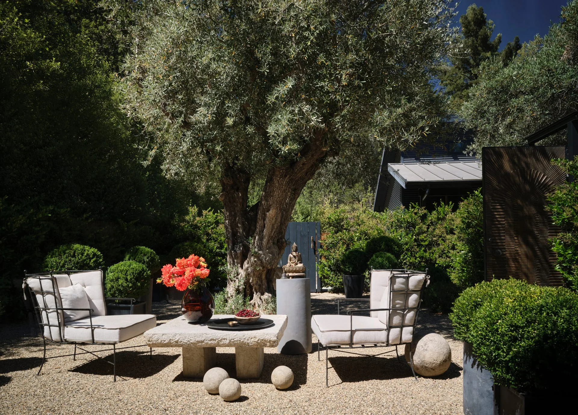 An outdoor gravel patio features two cushioned iron armchairs and a low stone coffee table arranged beneath a large olive tree.