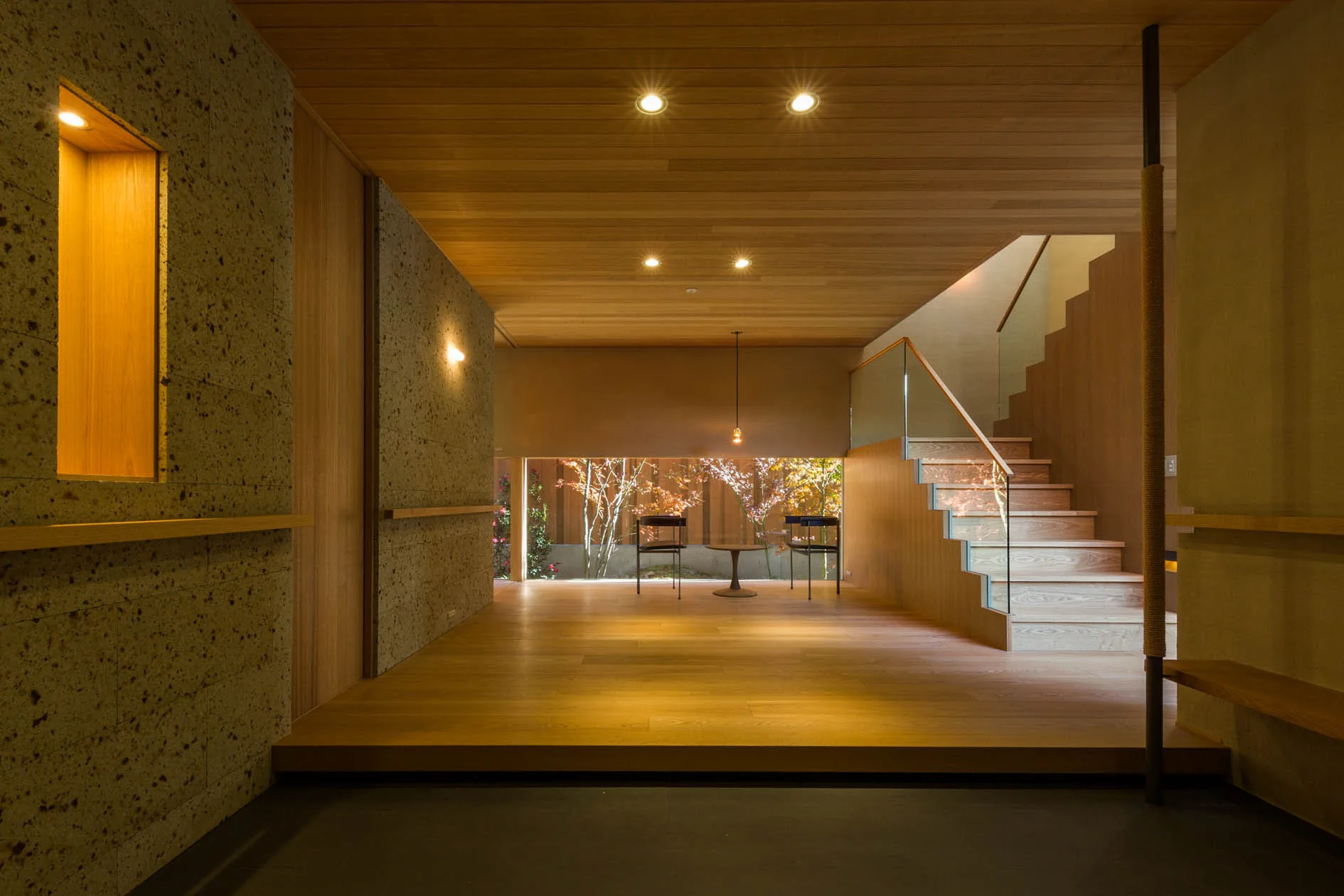 A minimalist wood hallway features a textured stone wall with a lit niche, a wooden staircase with a glass railing, and a low window showing a garden.