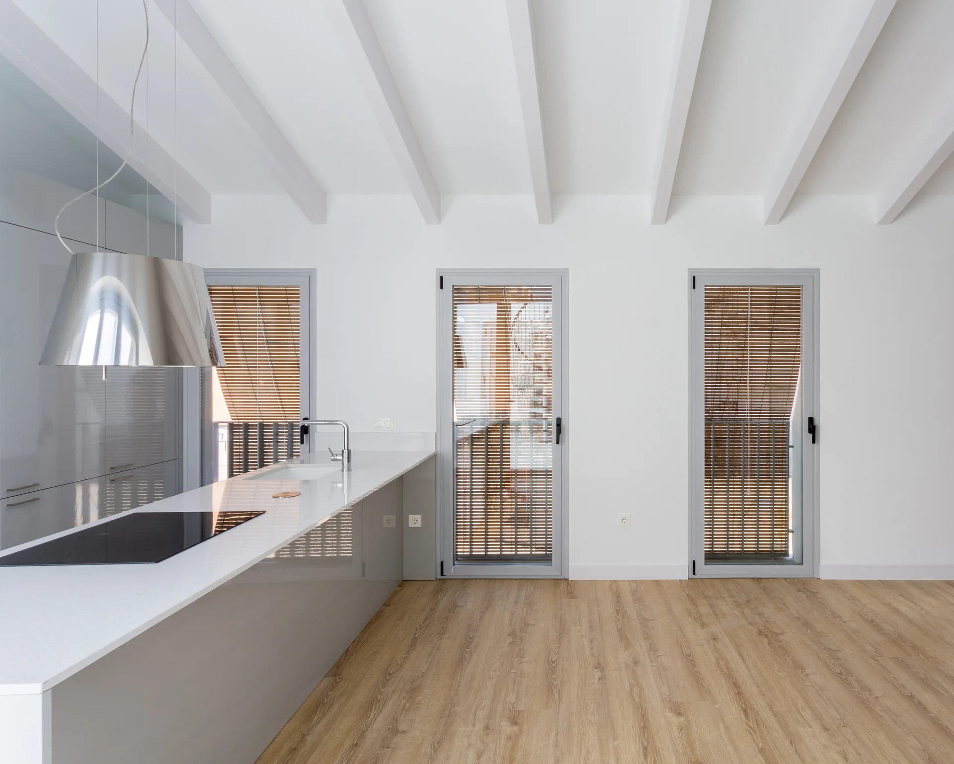 A minimalist kitchen features a glossy grey island, white quartz countertop, exposed white ceiling beams, and three glass doors with wood blinds.