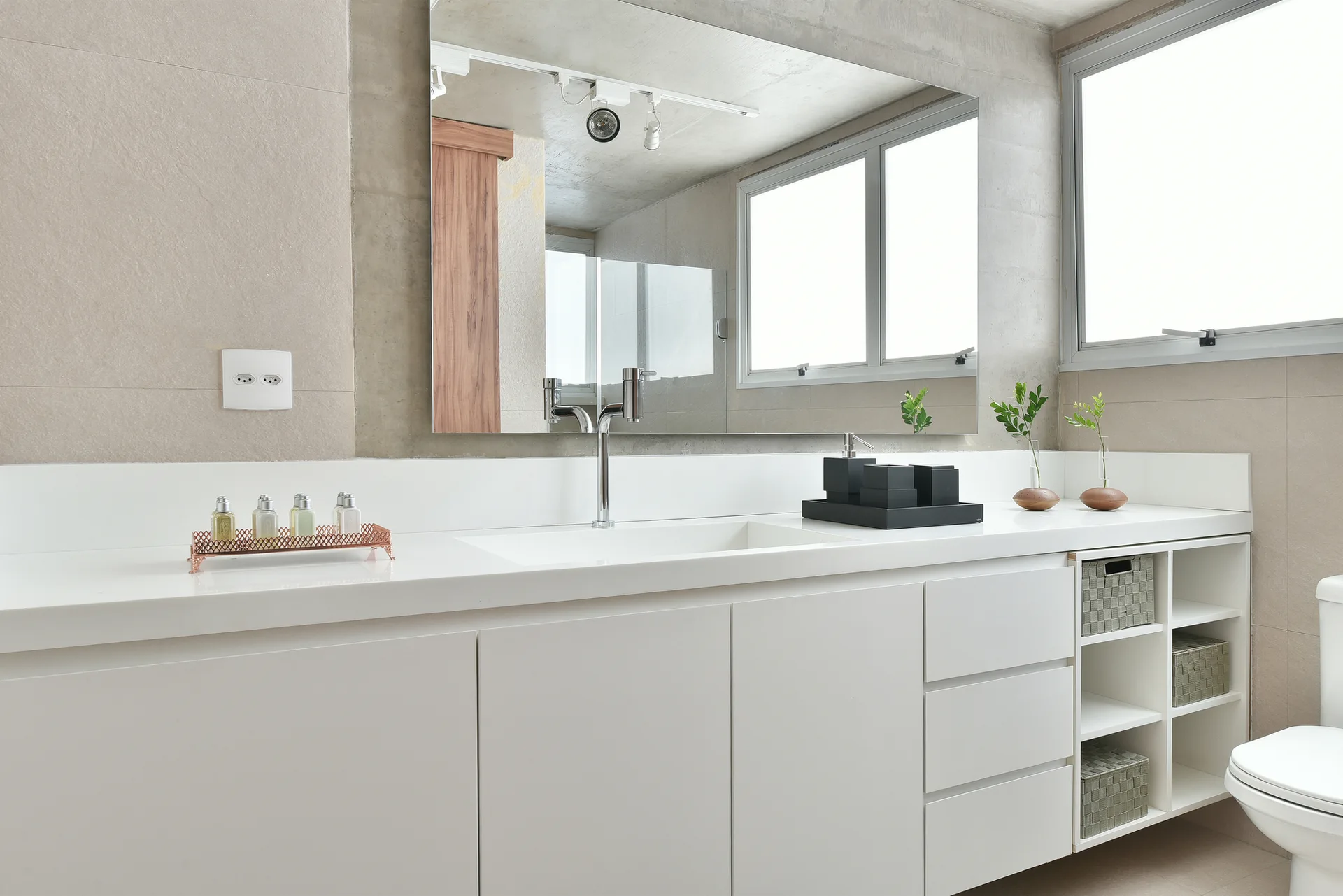 A modern bathroom features a white vanity with an integrated sink, a large rectangular wall mirror, and open storage cubbies with woven baskets.