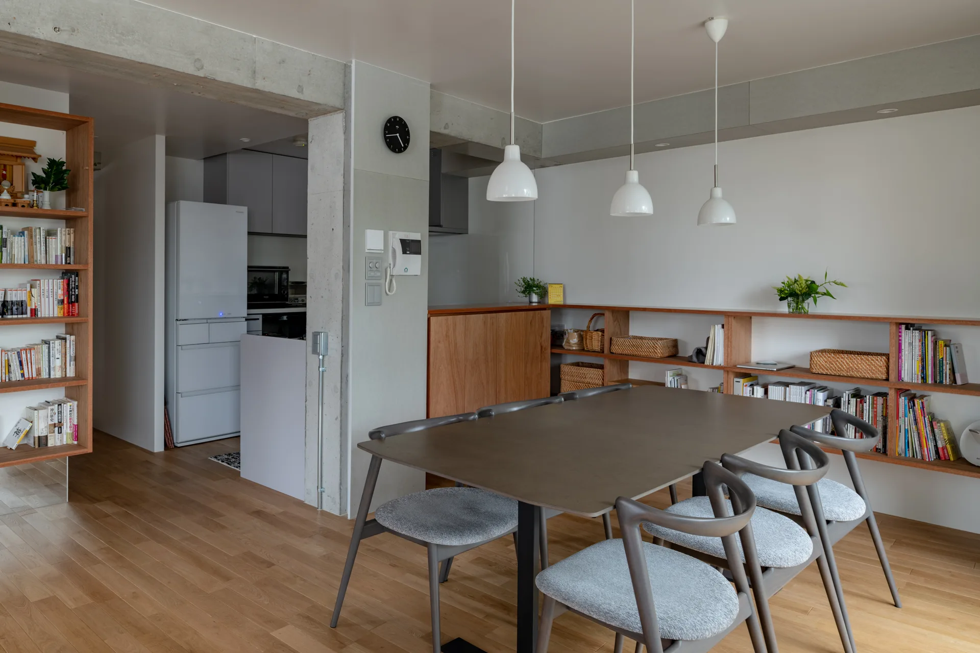 A modern dining room features a dark wood table, grey upholstered chairs, three white pendant lights, and a low built-in wooden bookshelf against a white wall.