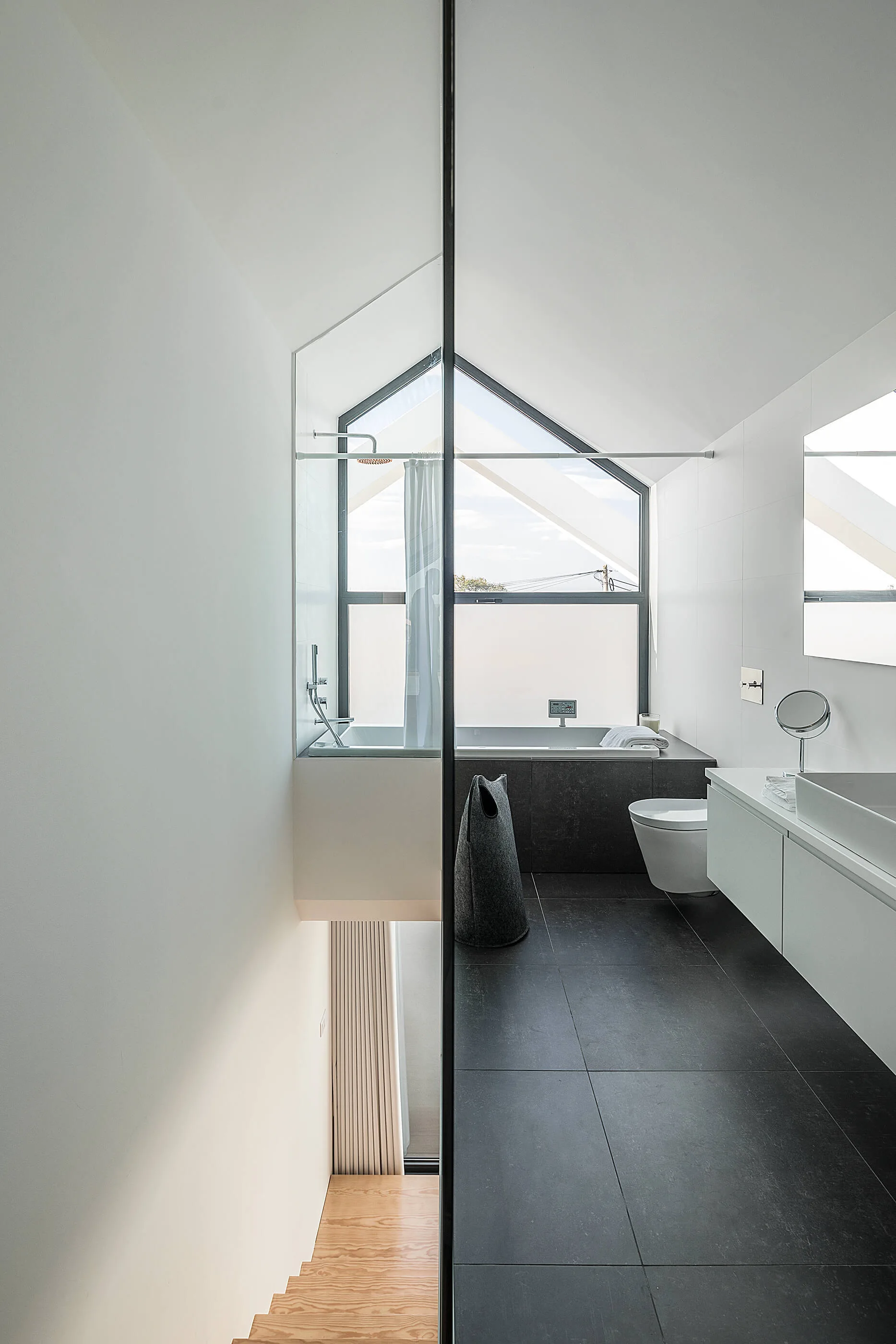 A minimalist bathroom with dark tiled floors features a white floating vanity, a bathtub under a pitched window, and a glass partition.