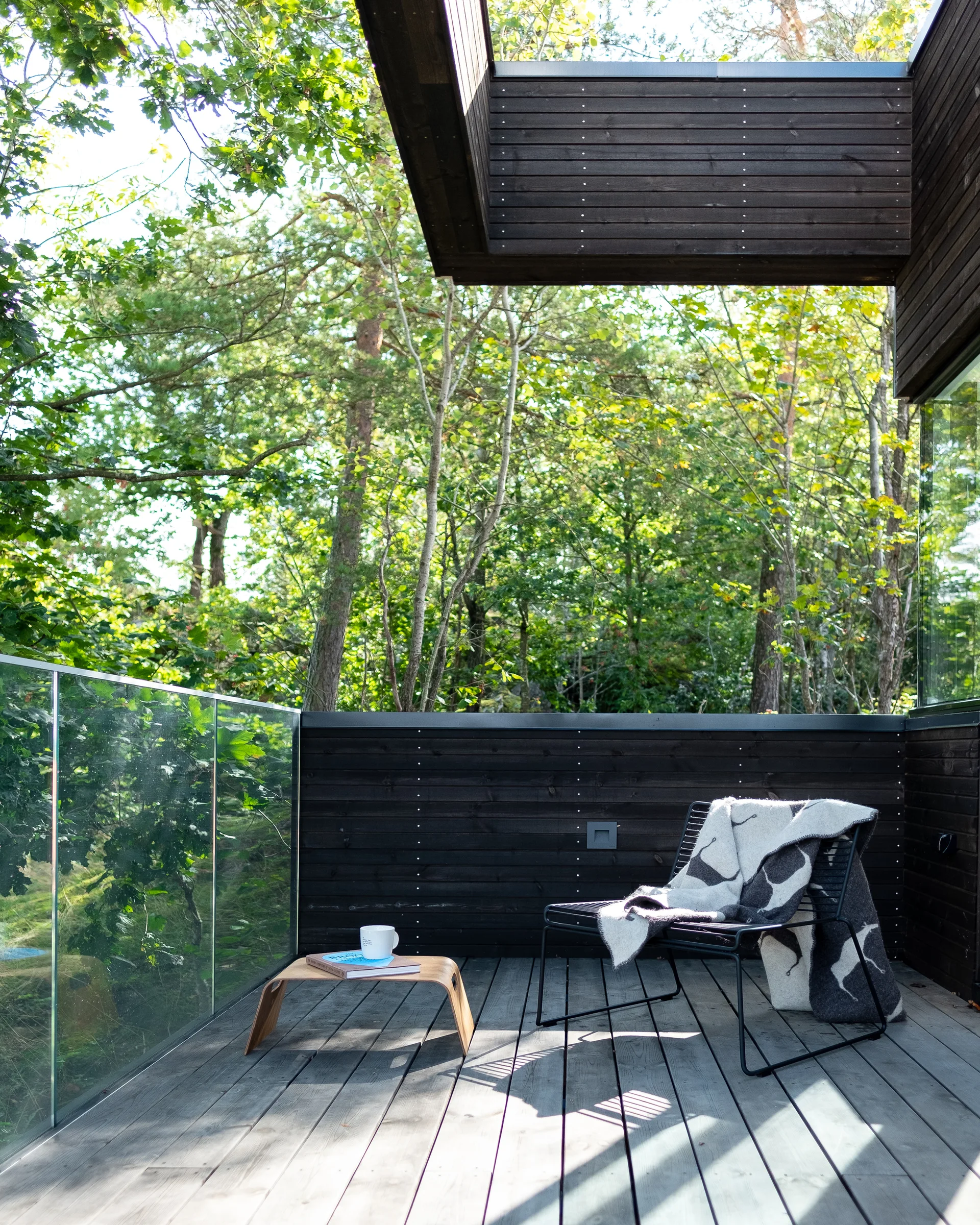 A modern balcony features dark wood siding, a glass railing, a black wireframe chair with a patterned throw, and a low wooden side table.
