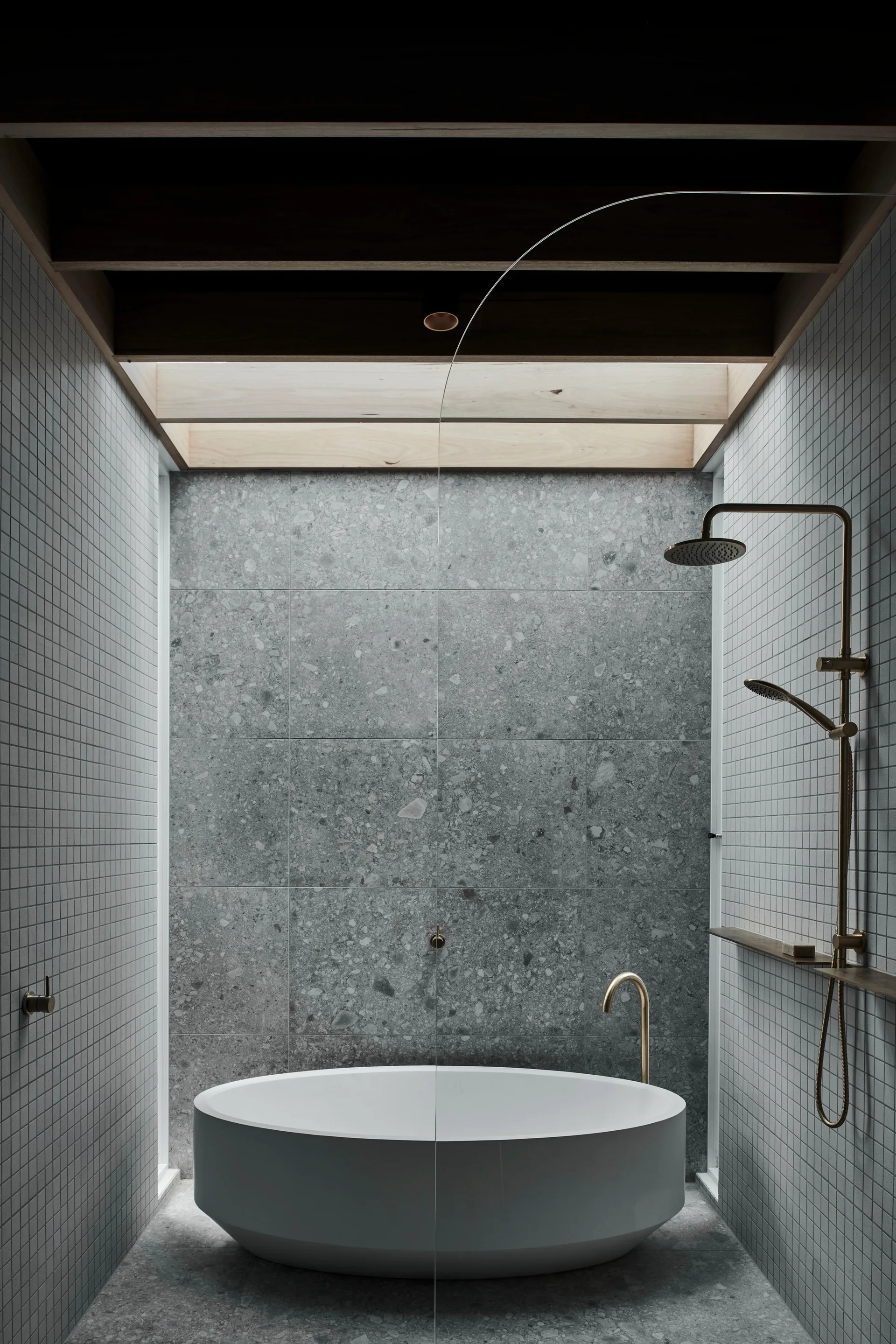A modern bathroom featuring a round freestanding white bathtub, grey terrazzo wall tiles, and bronze shower fixtures under a wood-framed skylight.