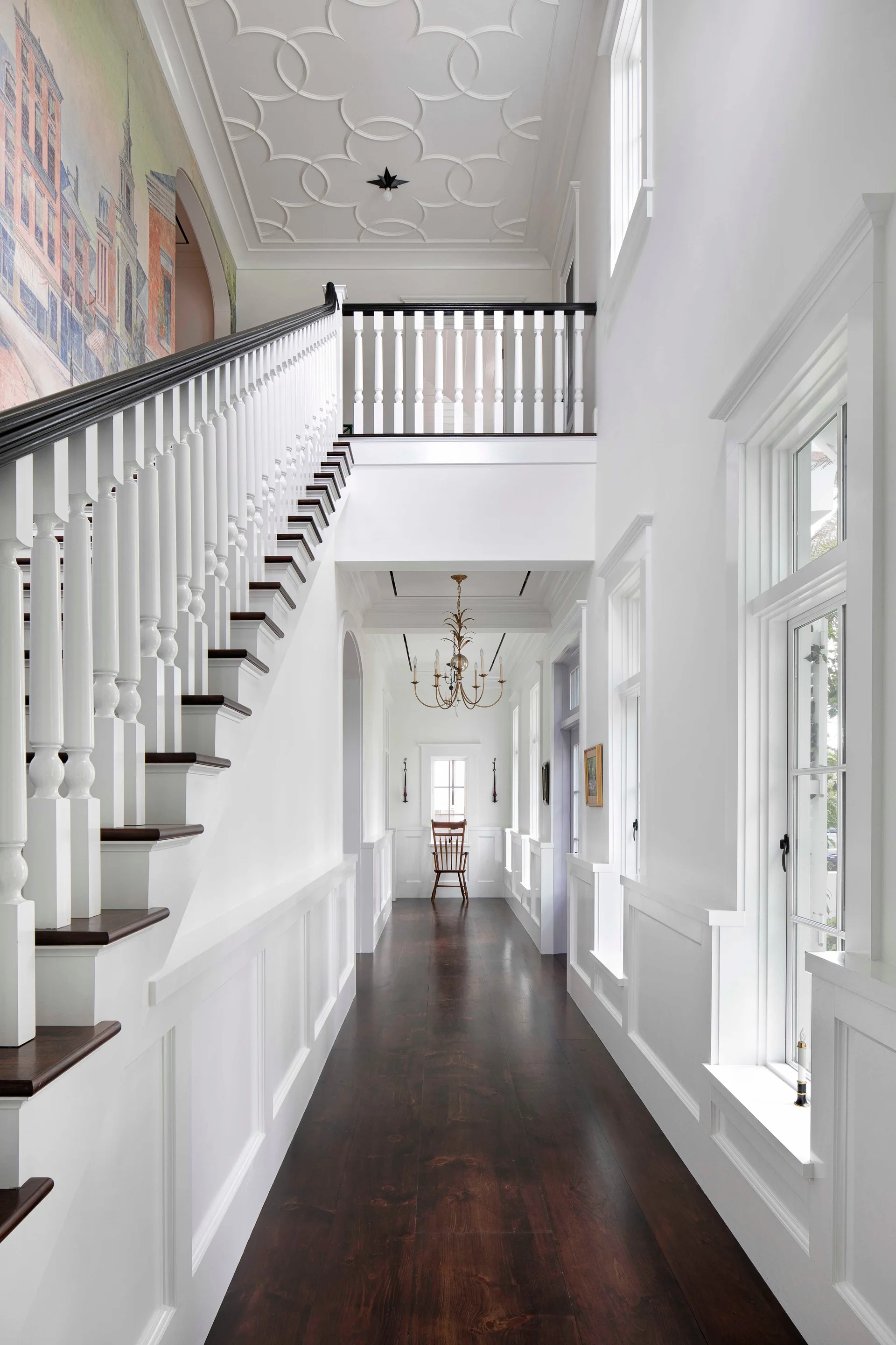 A bright white hallway features dark wood floors, a grand staircase with white railings, wainscoting, and a geometric plaster pattern on the ceiling.