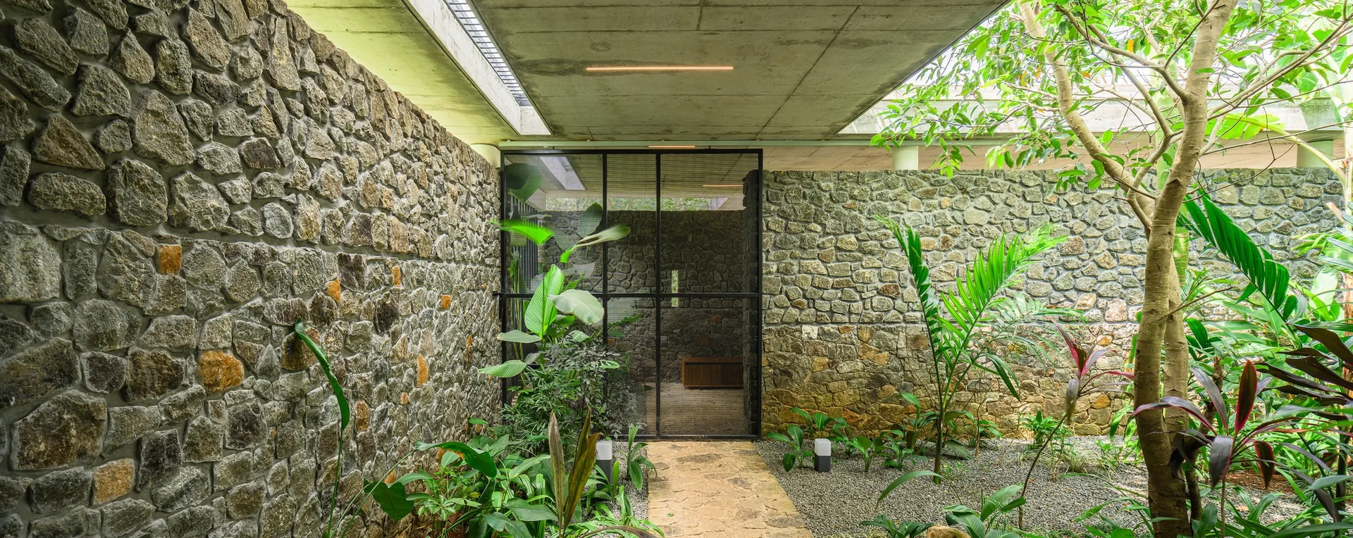 A semi-enclosed modern courtyard featuring rugged stone walls, a smooth concrete ceiling, a stone pathway, and lush green tropical plants.