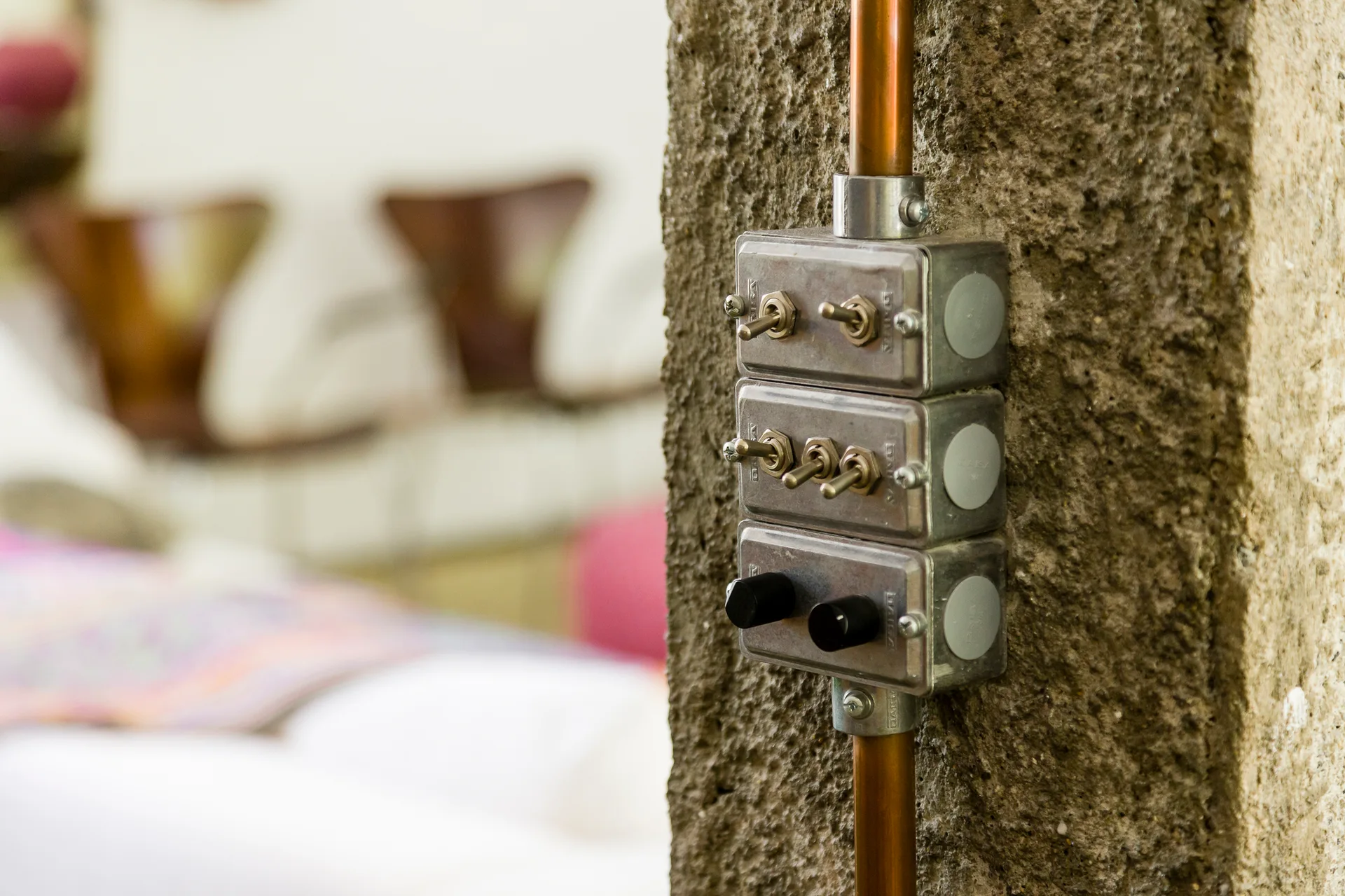 Three stacked industrial metal toggle switches and a copper conduit pipe are mounted on a rough, textured brown stone wall.