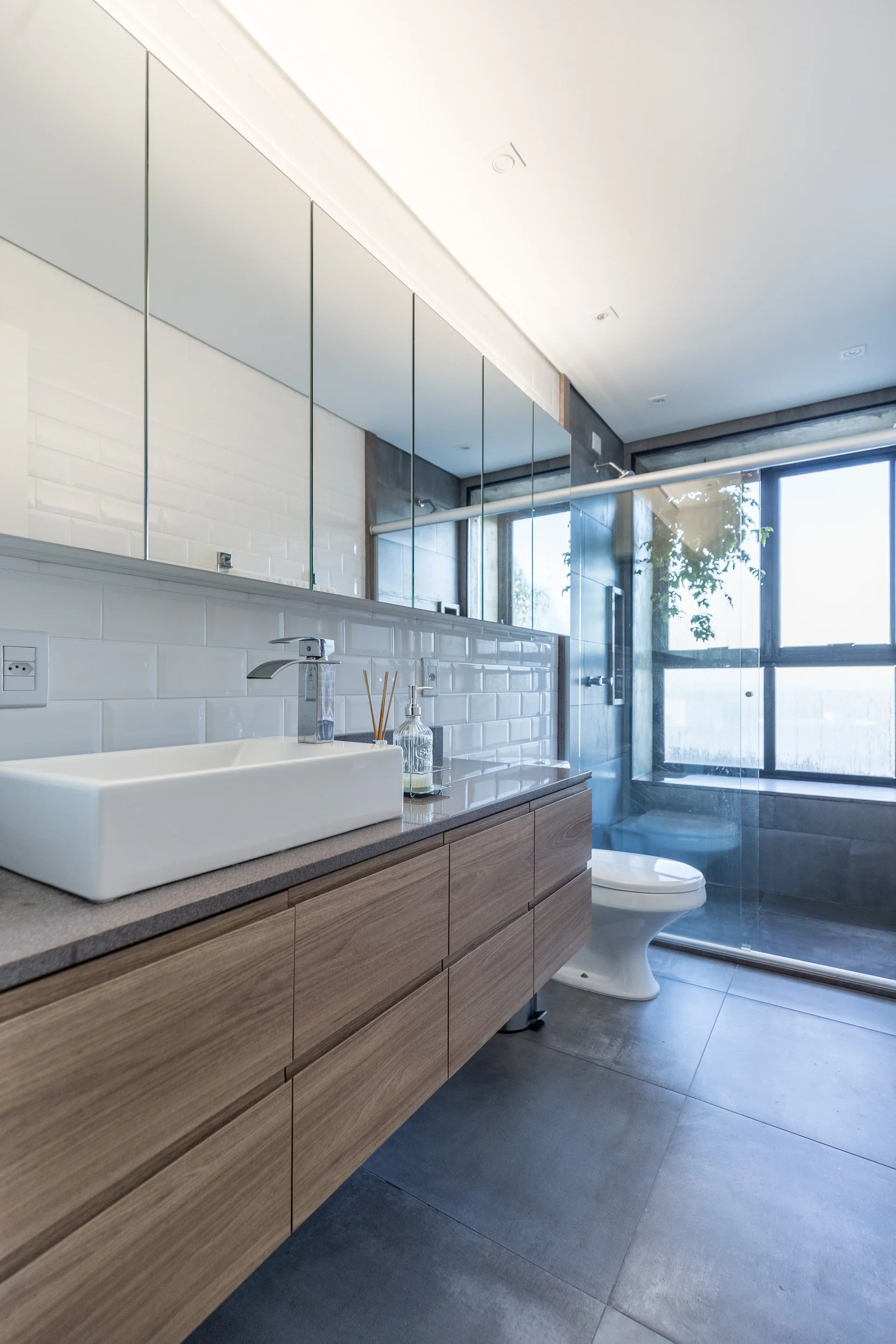A modern bathroom with a wooden floating vanity, a white vessel sink, and a large mirrored medicine cabinet against a white subway tile wall.