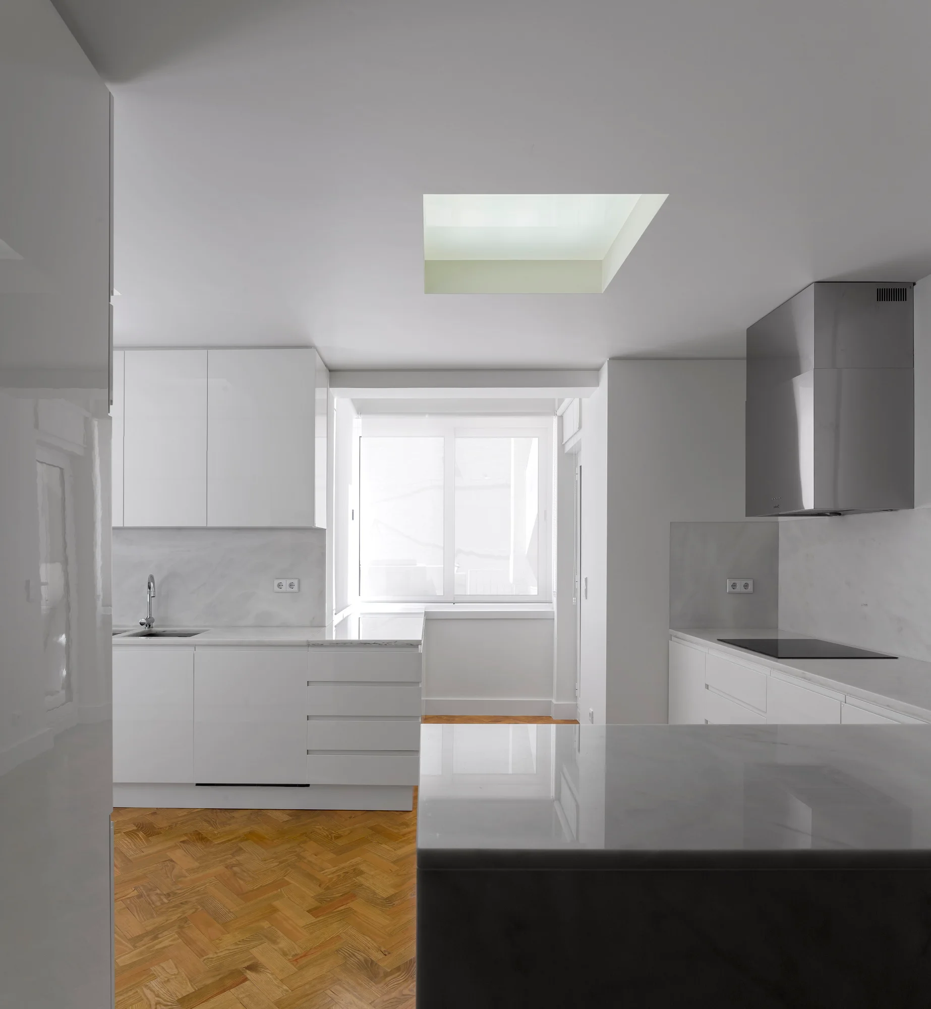 A minimalist white kitchen features flat-panel cabinets, marble countertops, a central skylight, and herringbone pattern wood flooring.