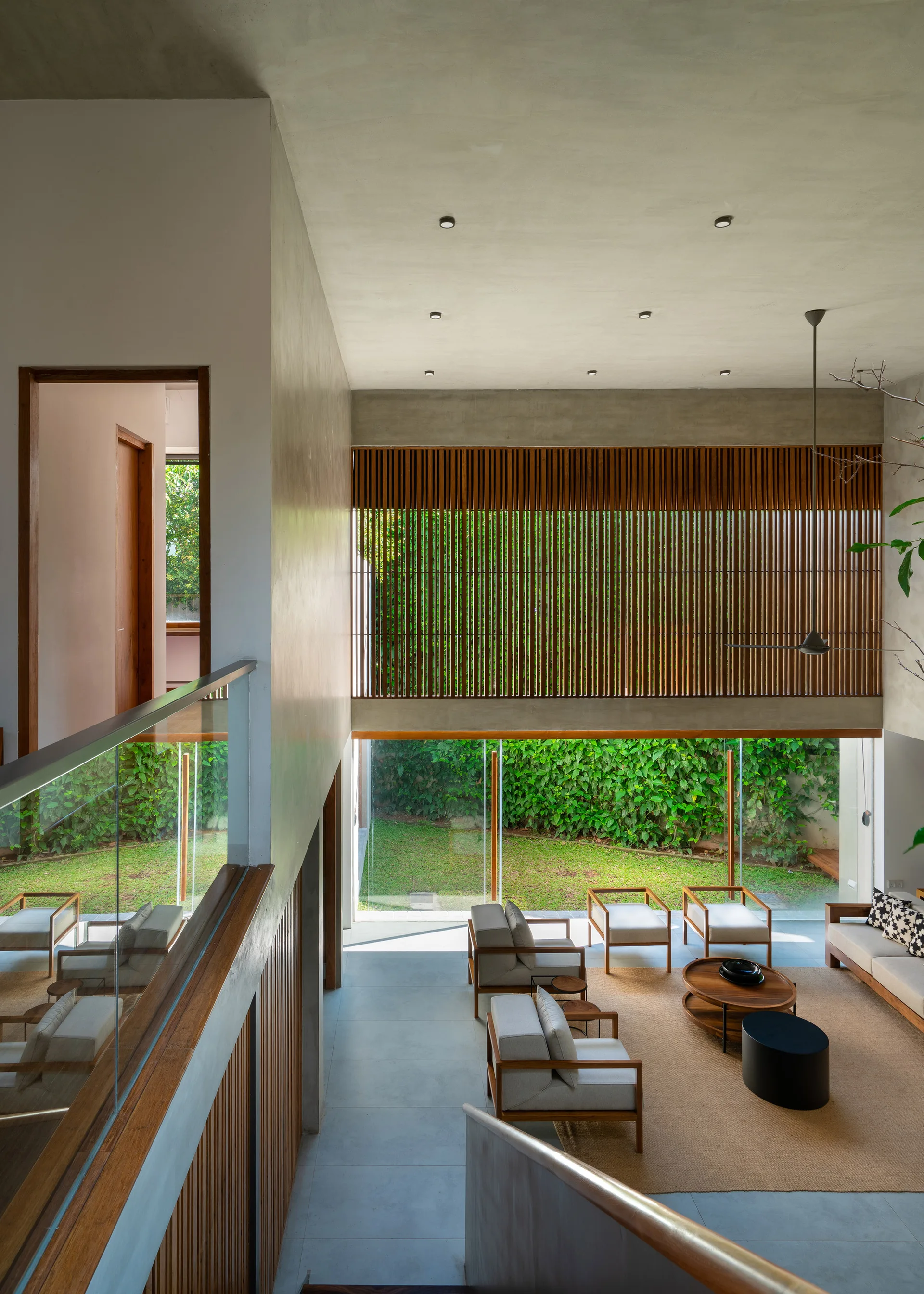 A view from a staircase looking down into a modern living room with wooden armchairs, concrete ceilings, and glass walls overlooking a green yard.