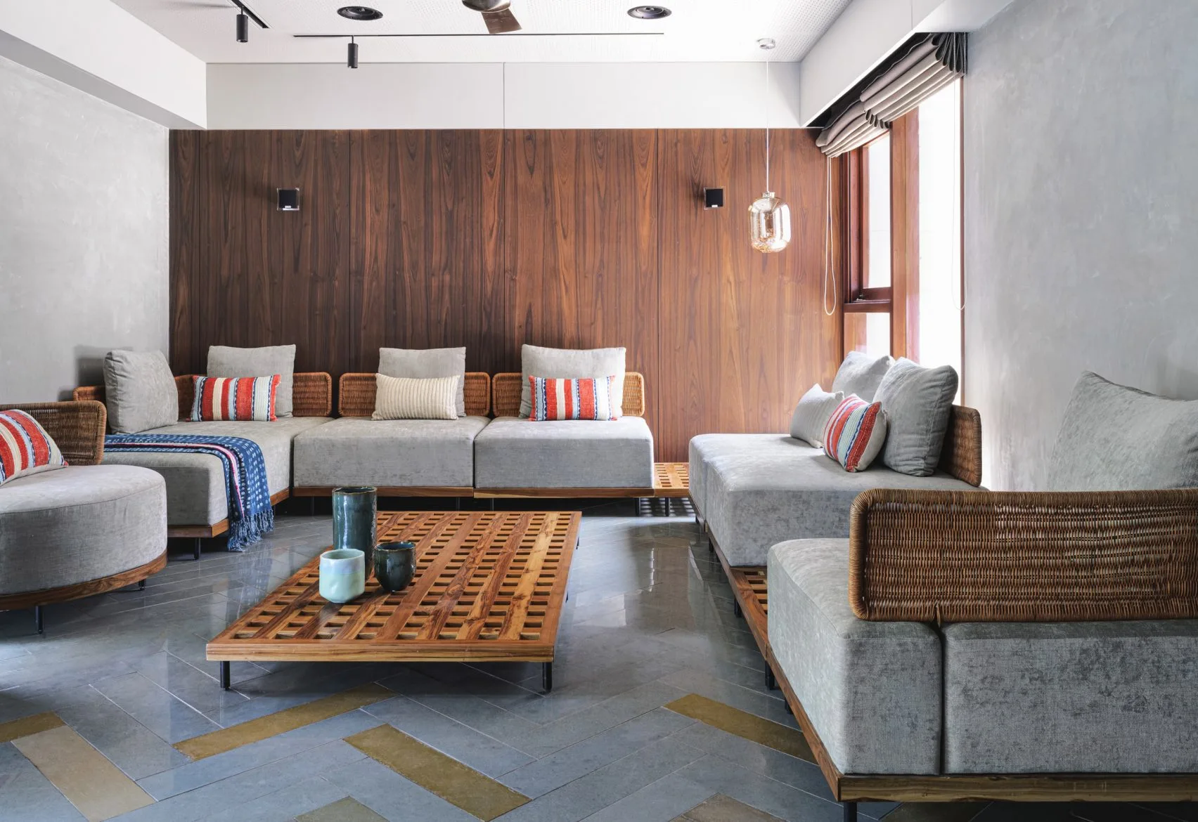 A lounge features a low modular grey sofa with woven rattan backing, a slatted wood coffee table, and a dark wood paneled wall.