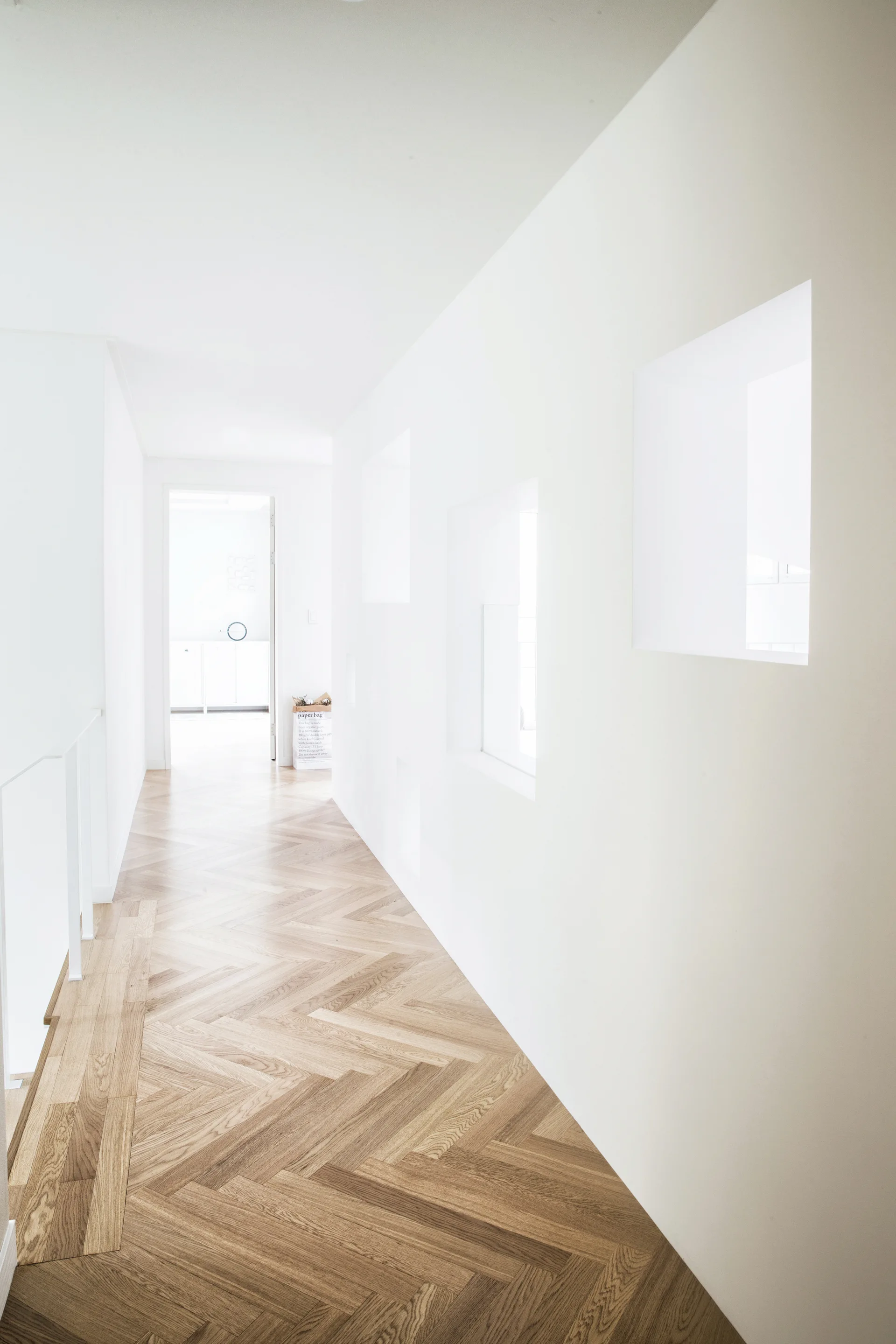 A bright minimalist hallway featuring light wood herringbone flooring, plain white walls, and a row of square architectural cutouts.