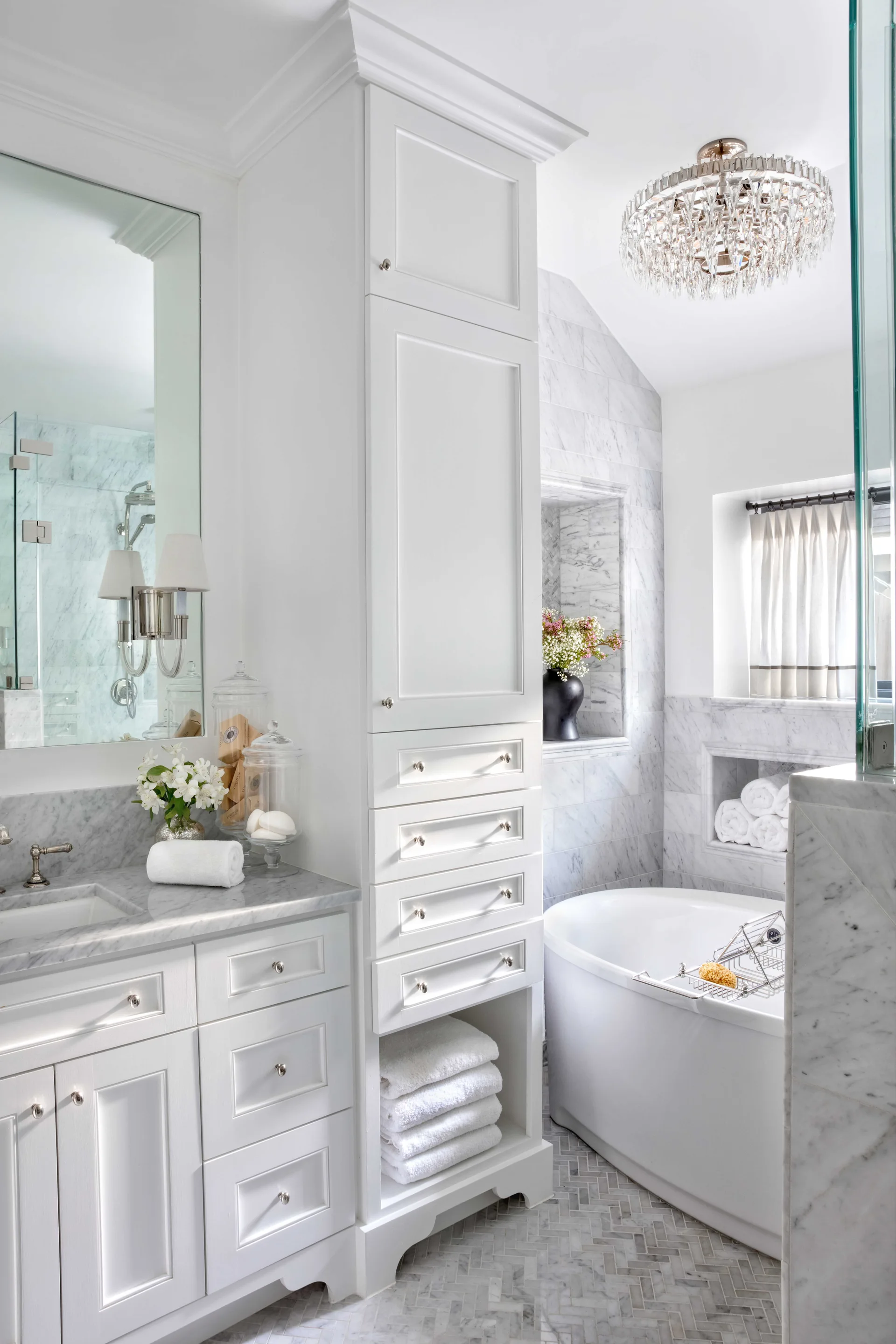 A white bathroom with a marble-topped vanity, a tall central linen cabinet, a freestanding tub, and herringbone floor tiles.