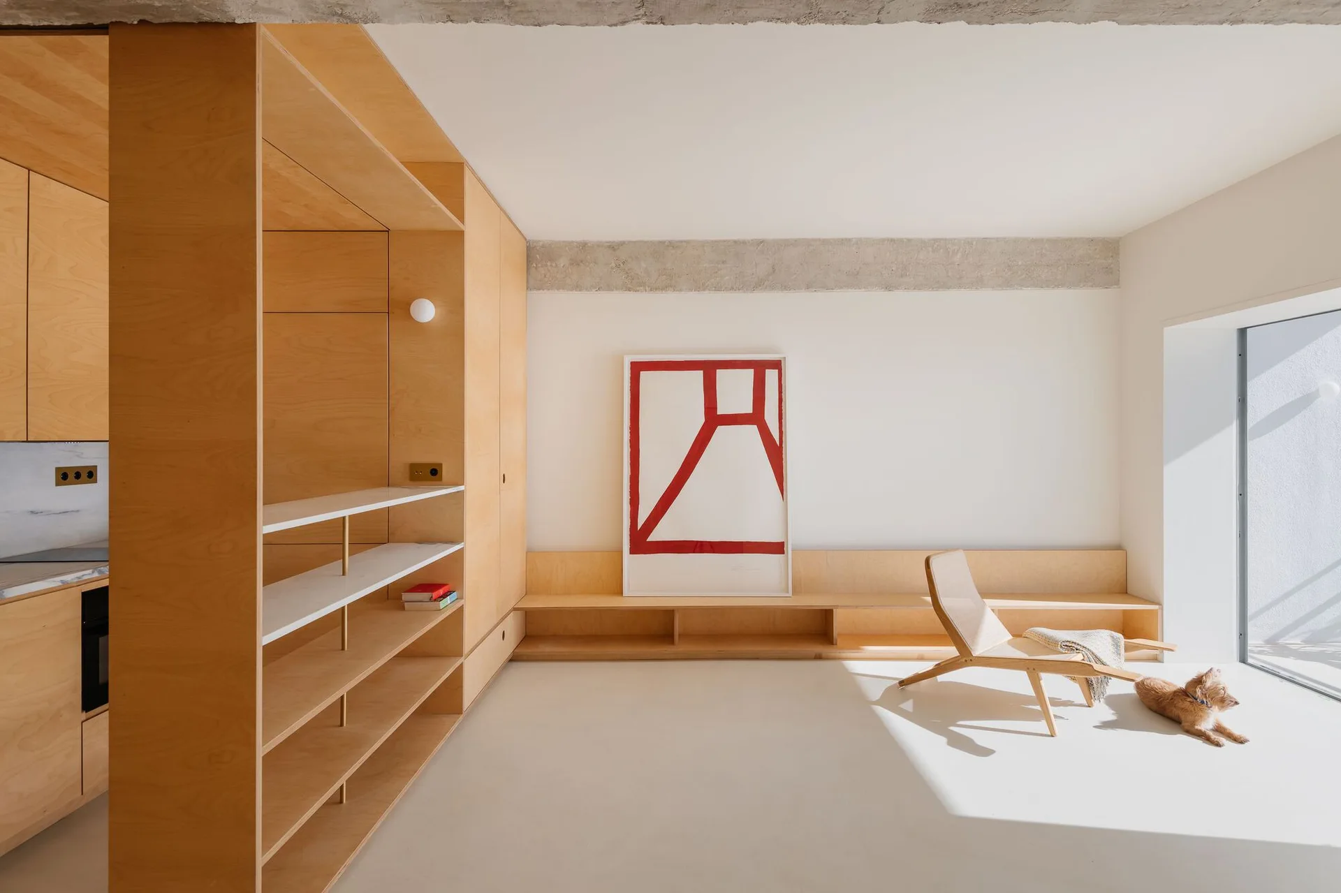 A minimalist living room features custom plywood cabinetry, a low built-in bench, an angular wooden lounge chair, and a large framed red graphic print.