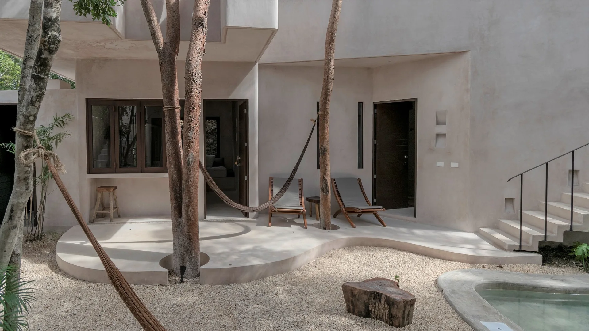 A beige plaster patio features a curved concrete slab with cutouts for mature trees holding a hammock, lounge chairs, and a small pool.