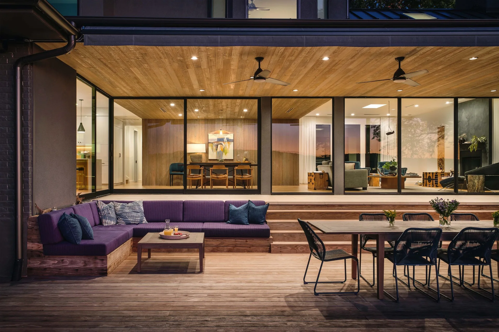A modern wood deck features a built-in purple sectional sofa, a matching coffee table, and an outdoor dining set under a wood-planked ceiling.
