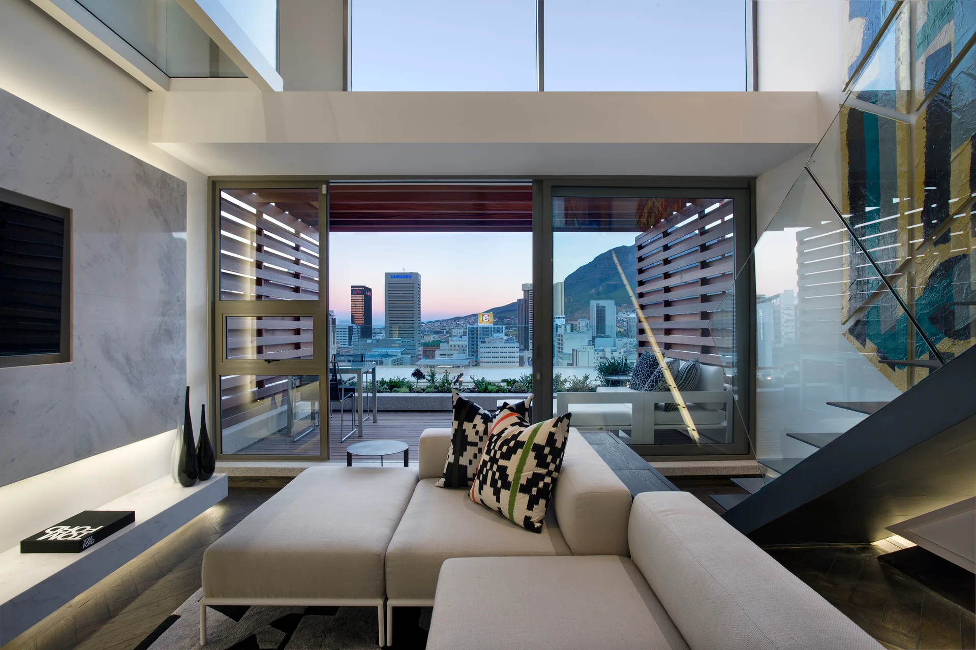A modern loft living room features a light gray sectional sofa, a glass staircase, and sliding doors opening to a wood-decked terrace with city views.