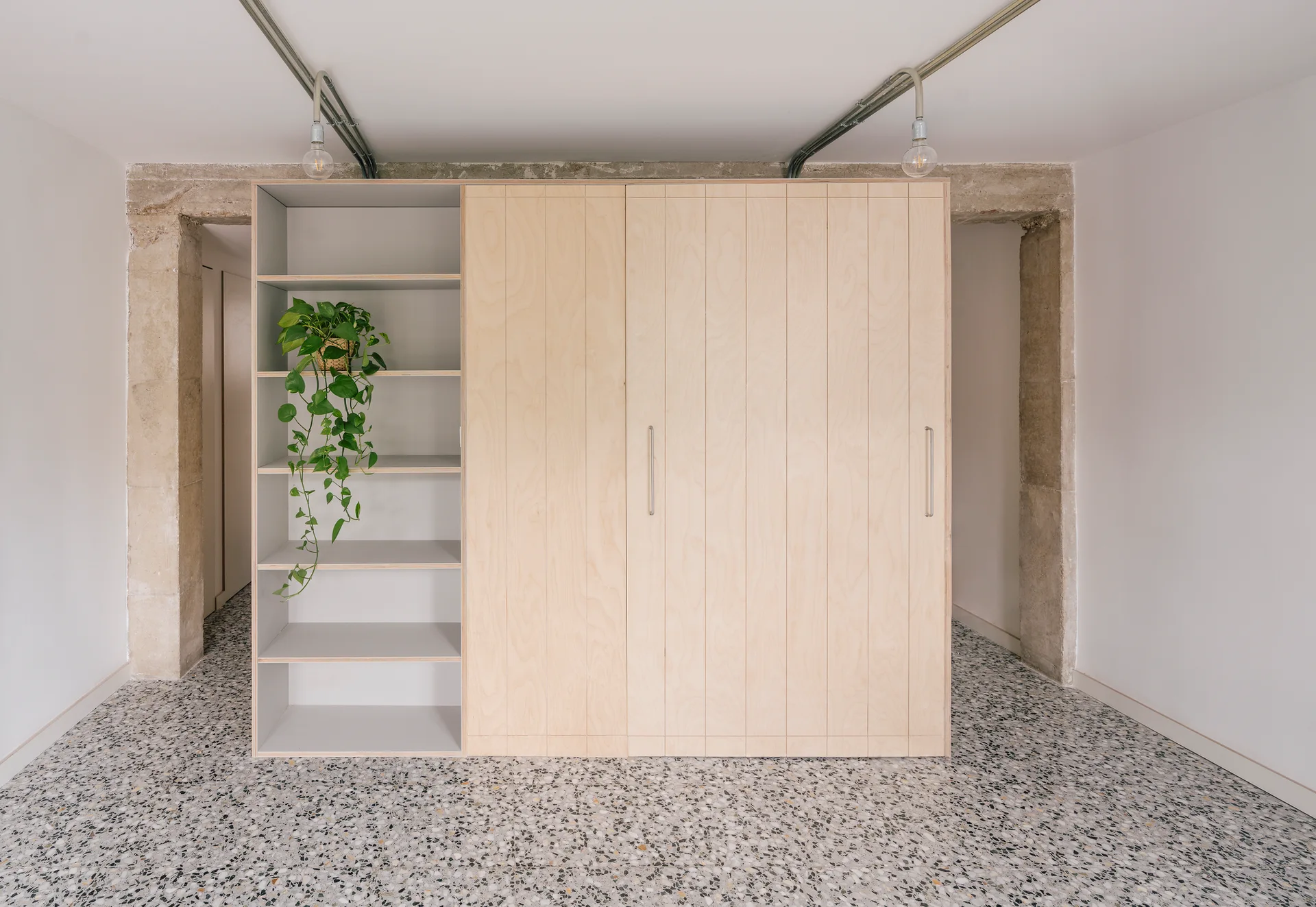 A minimalist plywood wardrobe with open shelving and sliding doors stands on a grey speckled terrazzo floor between raw stone entryways.