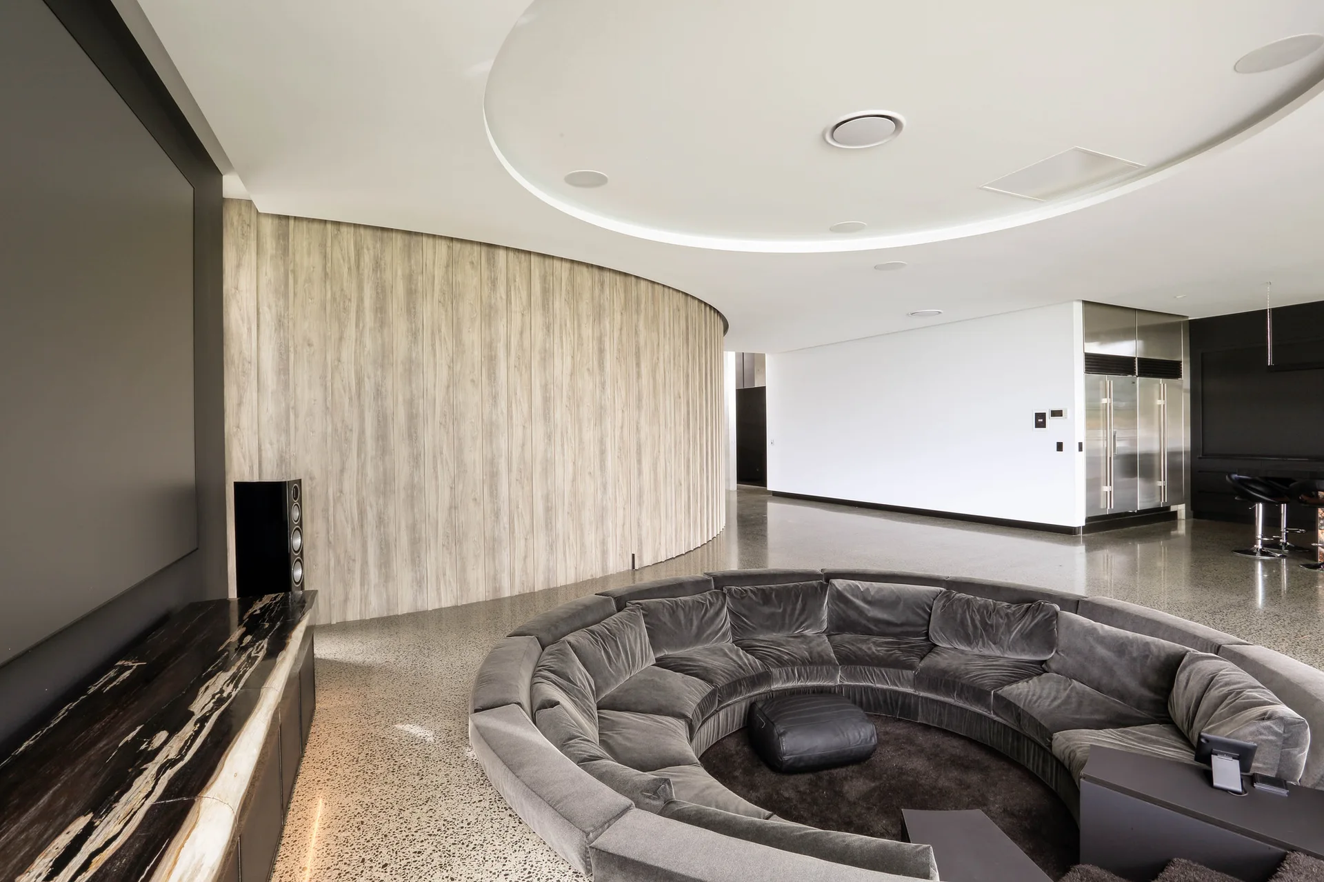 A large circular grey velvet sectional sofa sits in a spacious living area featuring a curved light wood paneled wall and circular ceiling cove lighting.
