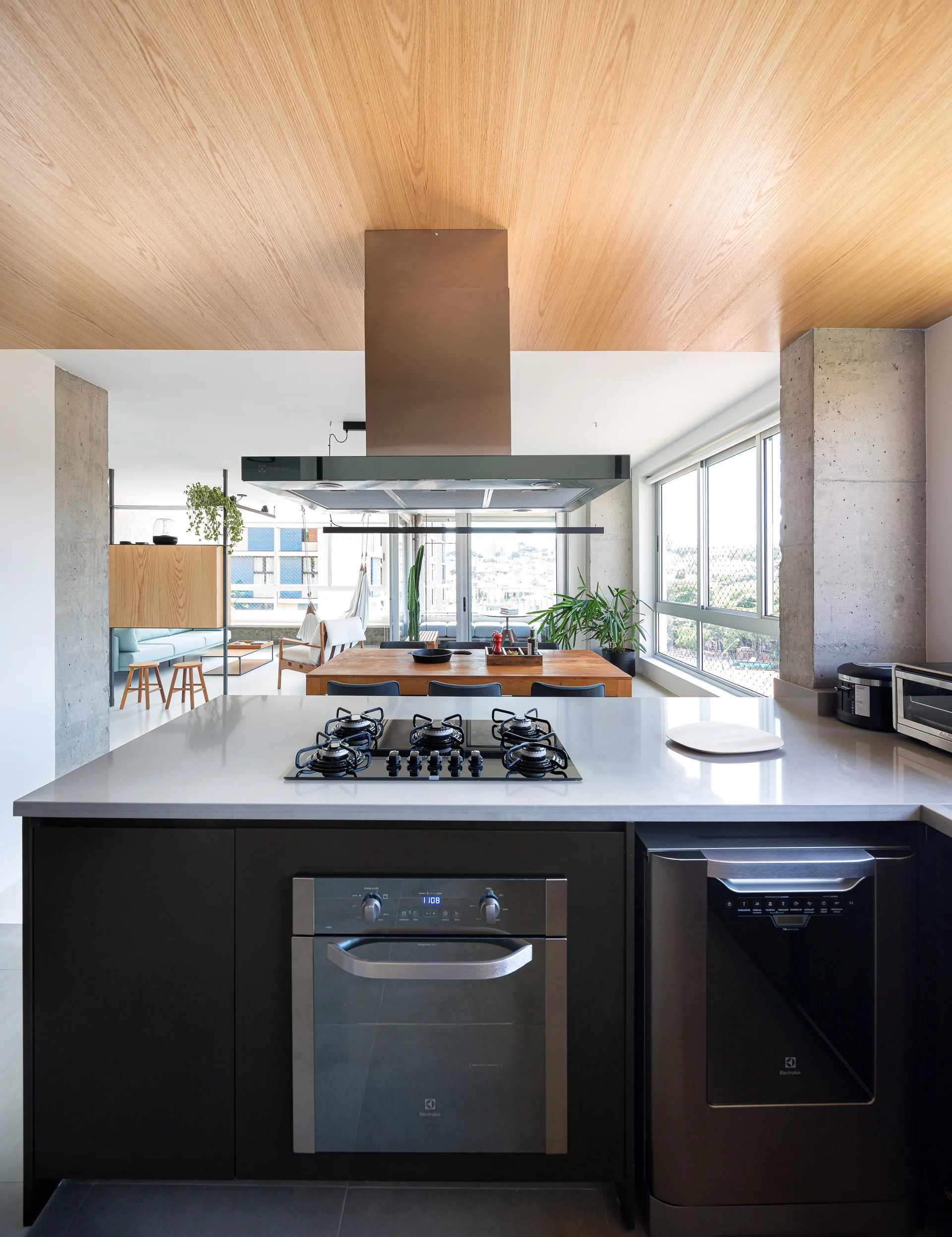 An open-plan kitchen featuring a dark kitchen island with a cooktop, a light wood ceiling panel, and exposed concrete pillars.