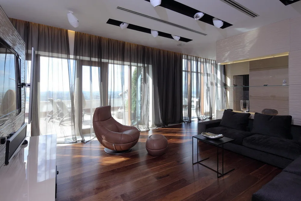 A contemporary living room featuring dark wood floors, a brown leather swivel chair, and large floor-to-ceiling windows with sheer dark curtains.