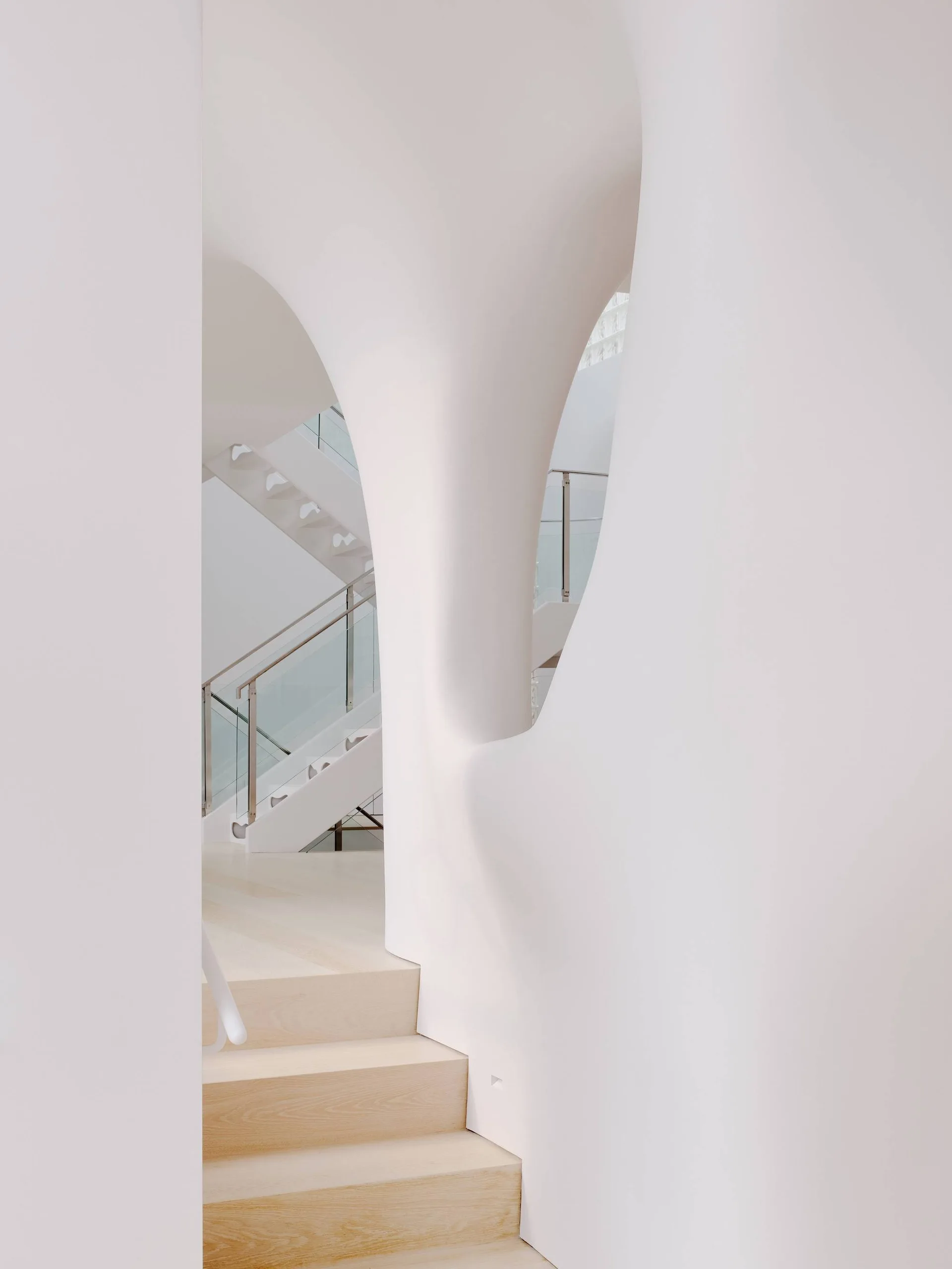 A contemporary staircase with light wood treads and glass railings is framed by a smooth, organic white sculptural plaster wall.