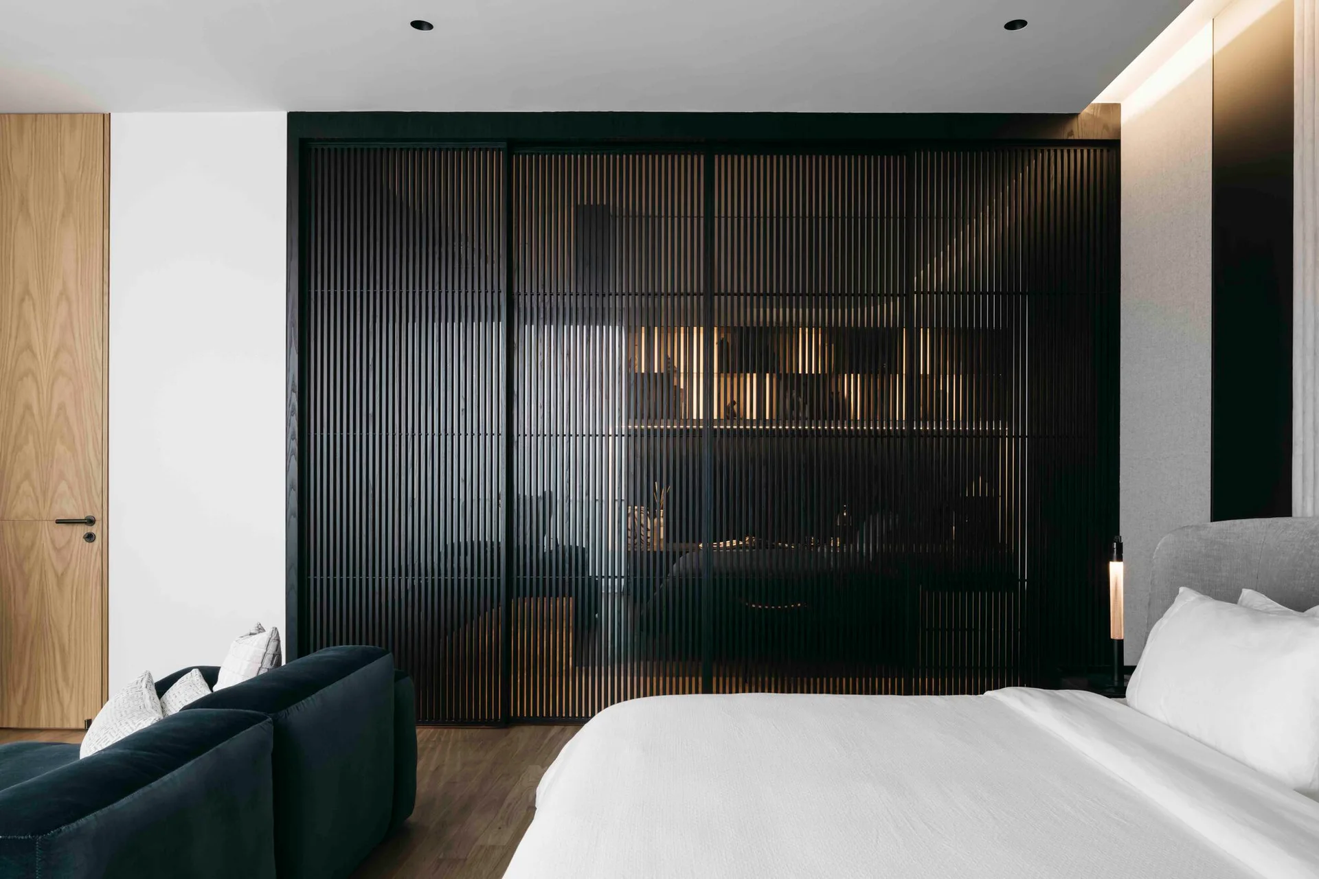 A modern bedroom featuring a bed with white linens next to a large black slatted sliding partition illuminated by warm light.