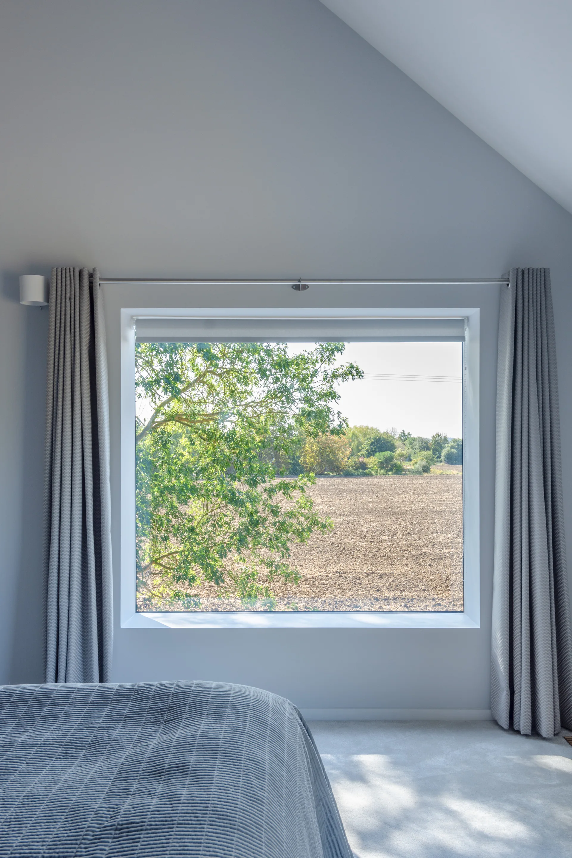 A minimalist bedroom featuring a large picture window framed by grey curtains, with a quilted bed in the foreground and a view of a green tree outside.