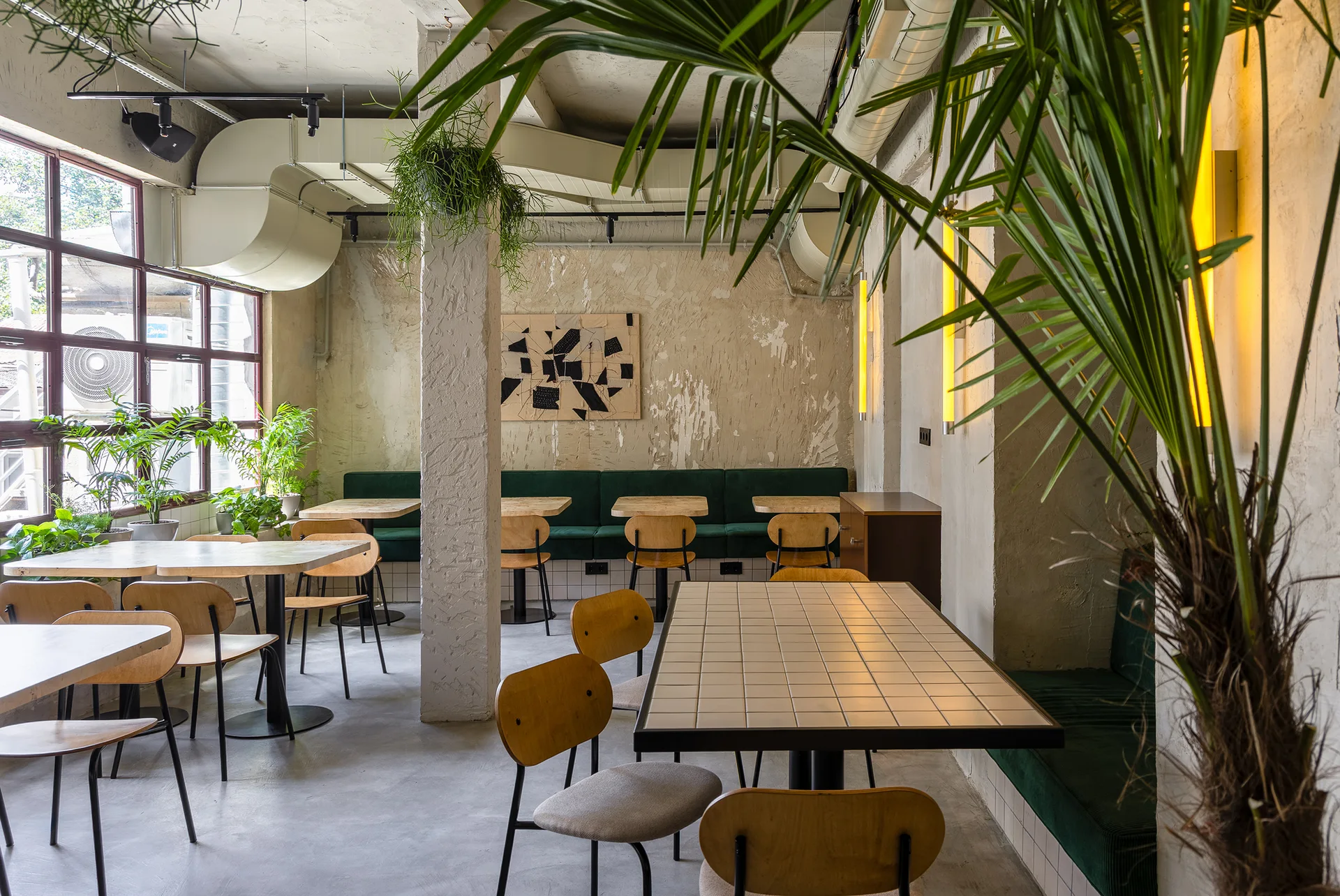 An industrial dining space with concrete floors, distressed plaster walls, wooden tables, green banquette seating, and exposed ceiling ductwork.