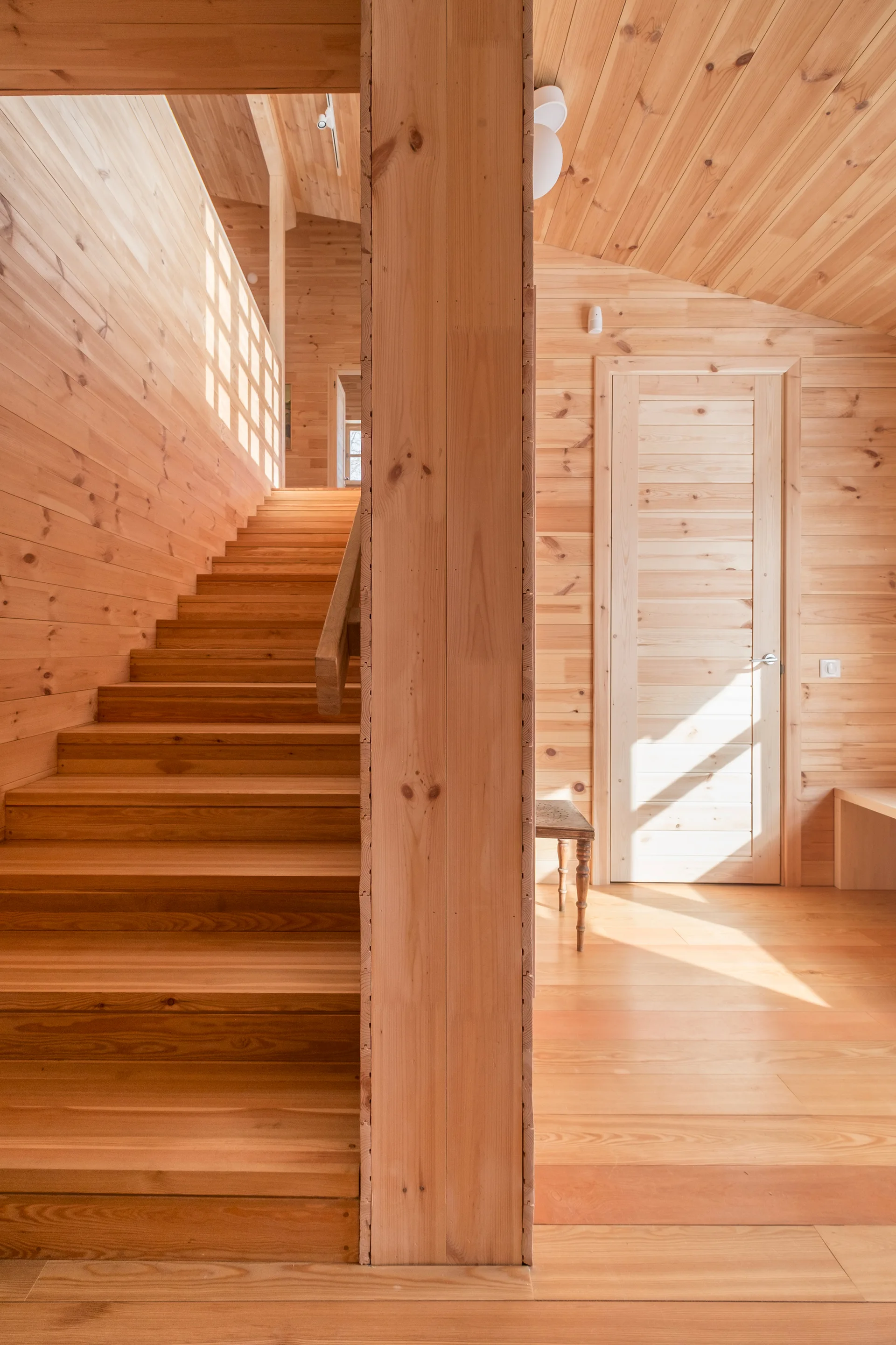 A residential hallway constructed entirely of light-toned pine wood features a prominent central staircase, sloped ceiling, and a matching wooden door.
