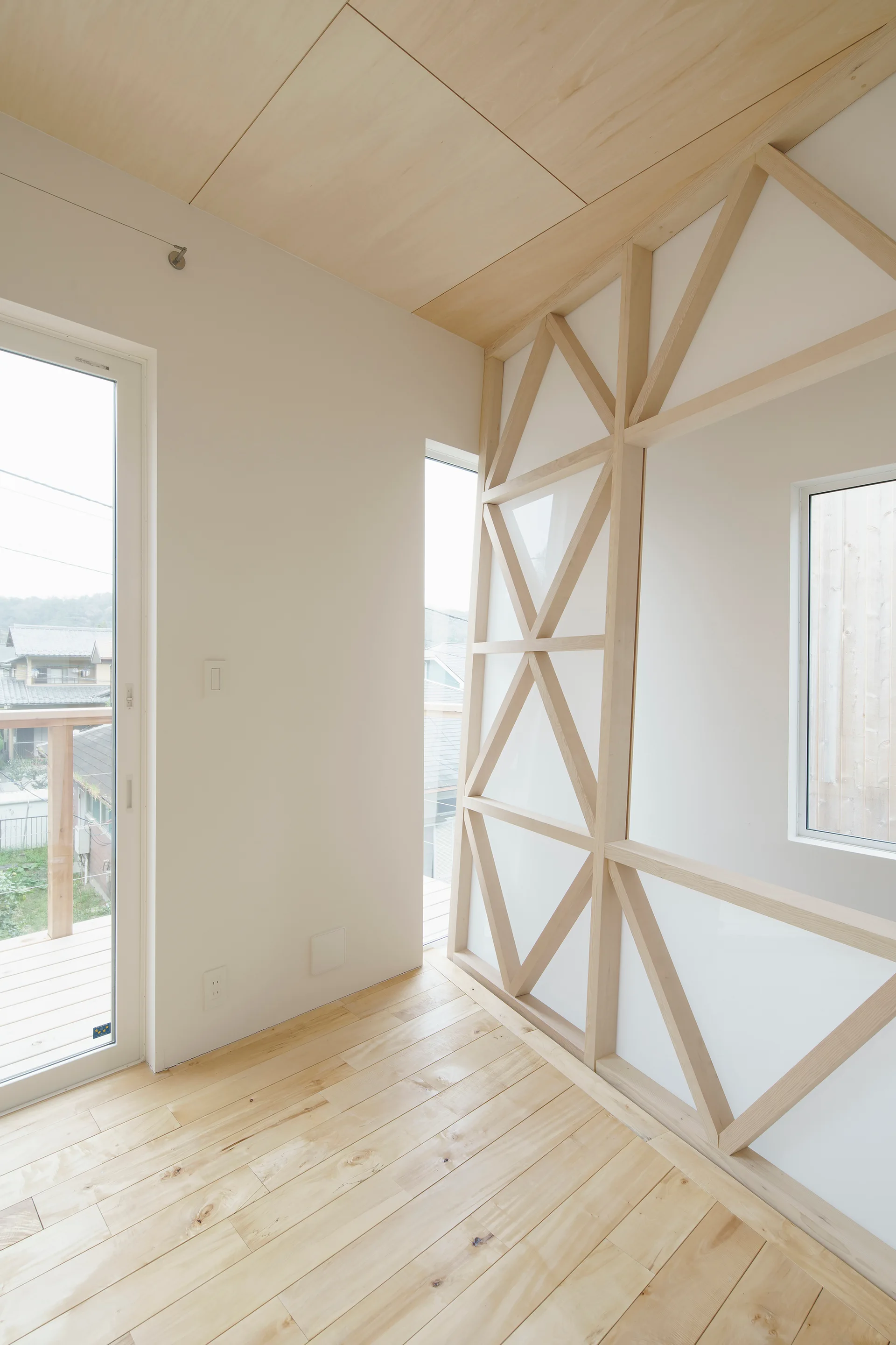An empty room featuring light wood flooring, a smooth plywood ceiling, and an exposed geometric timber frame on a white wall next to tall windows.
