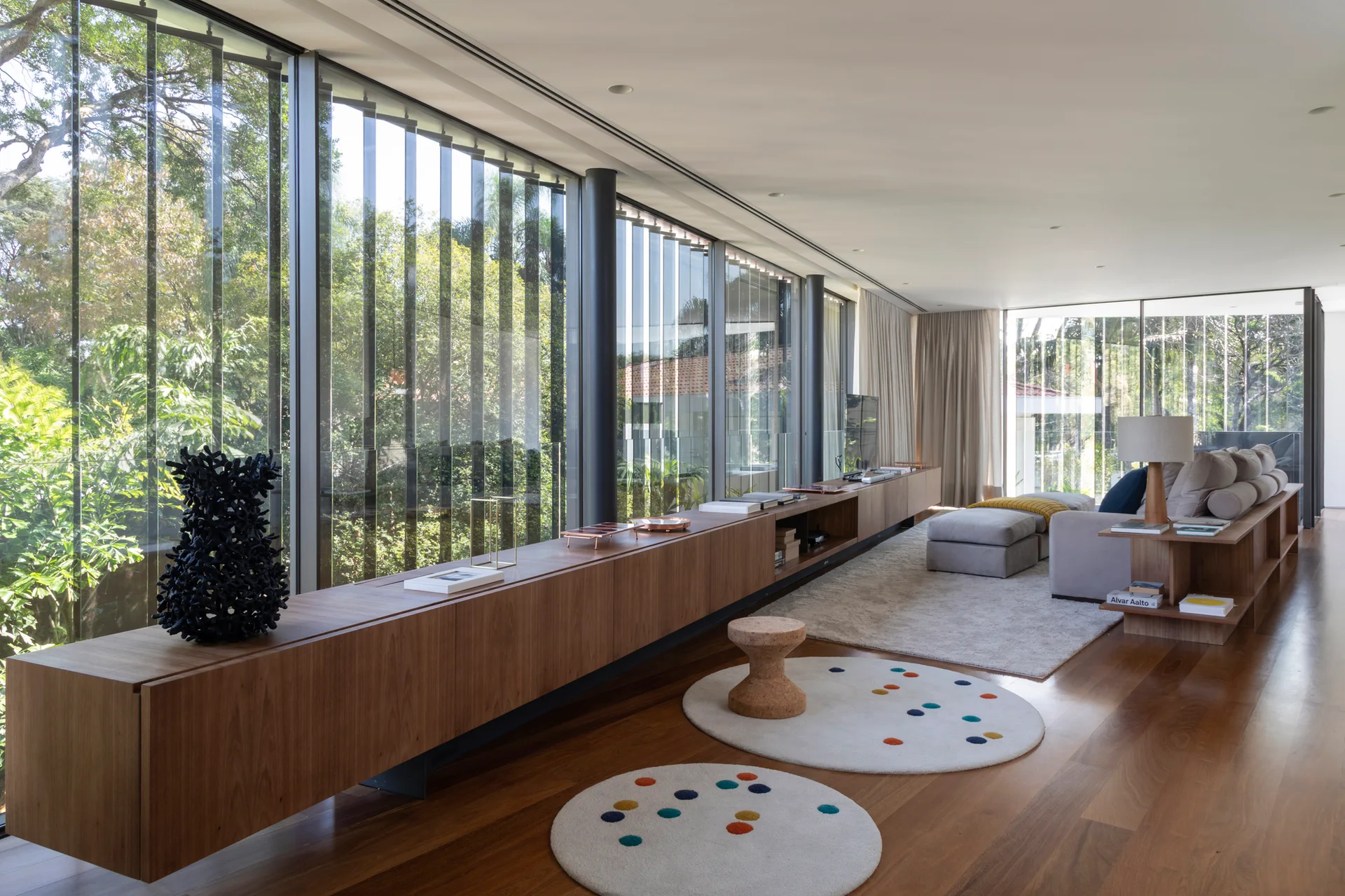 A modern living room features a long floating wood console alongside floor-to-ceiling windows, with a neutral sofa and two dotted round rugs.
