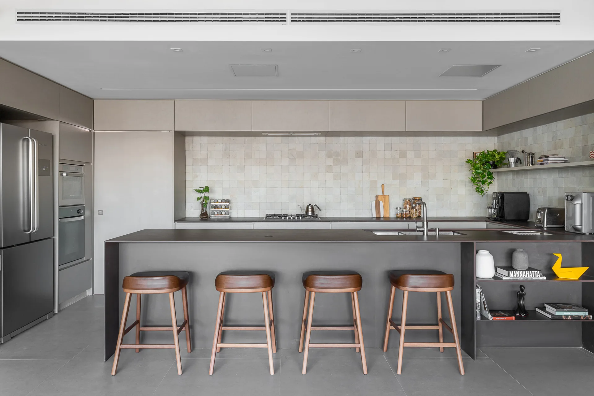 A minimalist kitchen features a long dark gray island with four wooden stools, light beige square tiled backsplash, and stainless steel appliances.