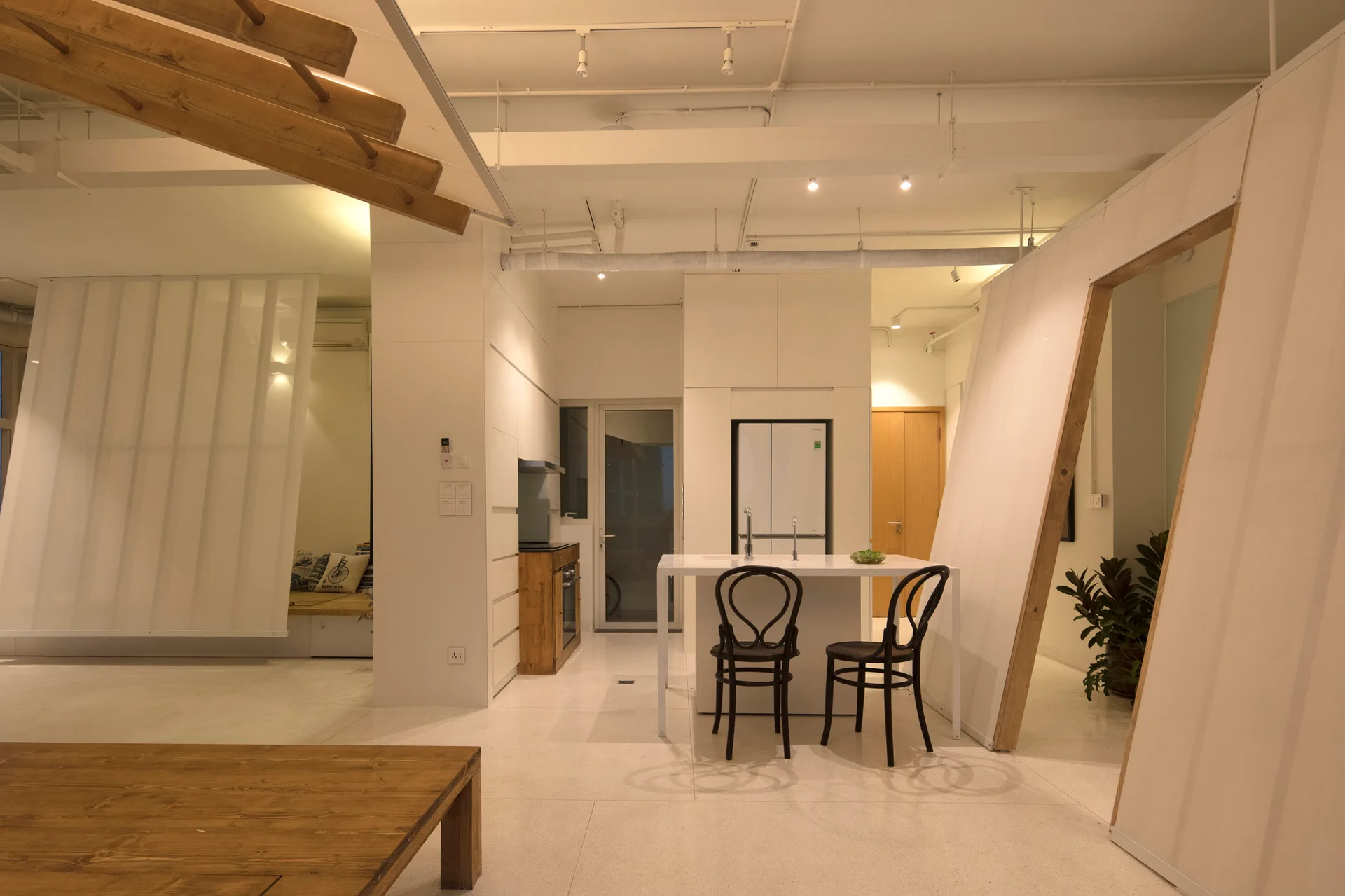 A modern white kitchen features a central island with two black bentwood chairs, surrounded by angled partition walls and an exposed white ceiling.