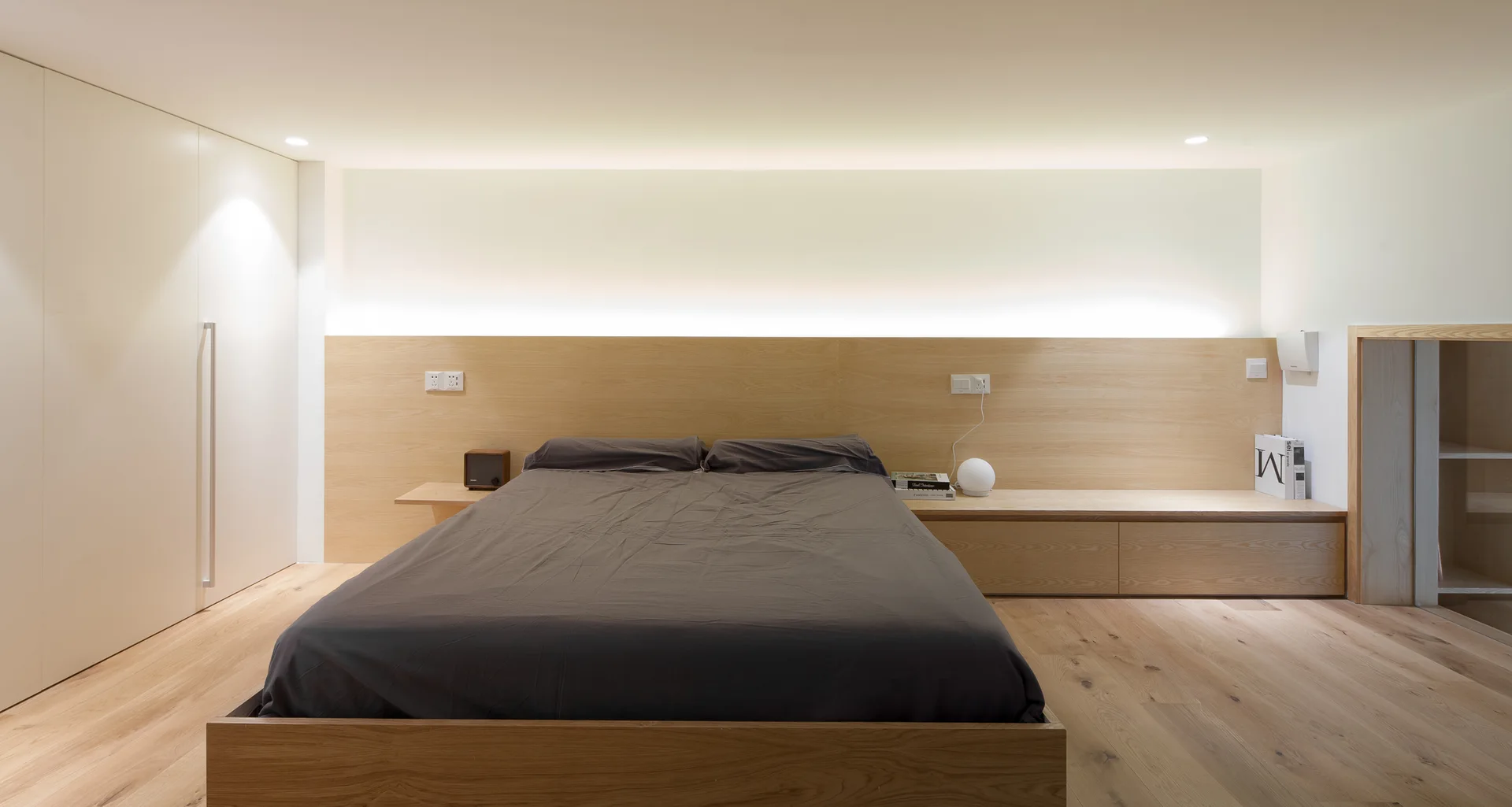 A minimalist bedroom featuring a grey platform bed against a light wood paneled wall with integrated bedside ledges and warm cove lighting.