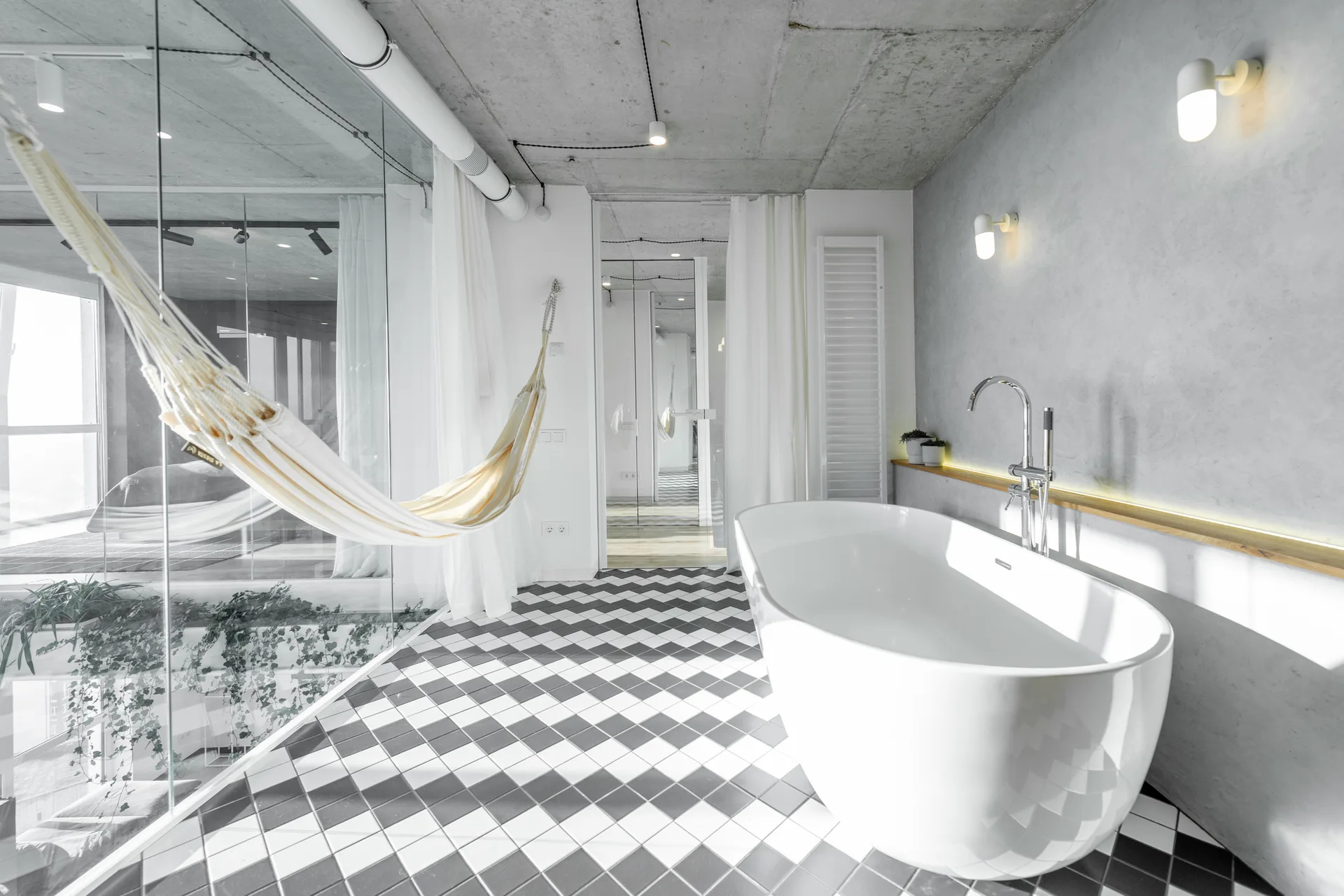 A modern bathroom features a white freestanding bathtub on a black and white checkered floor beneath a raw concrete ceiling.