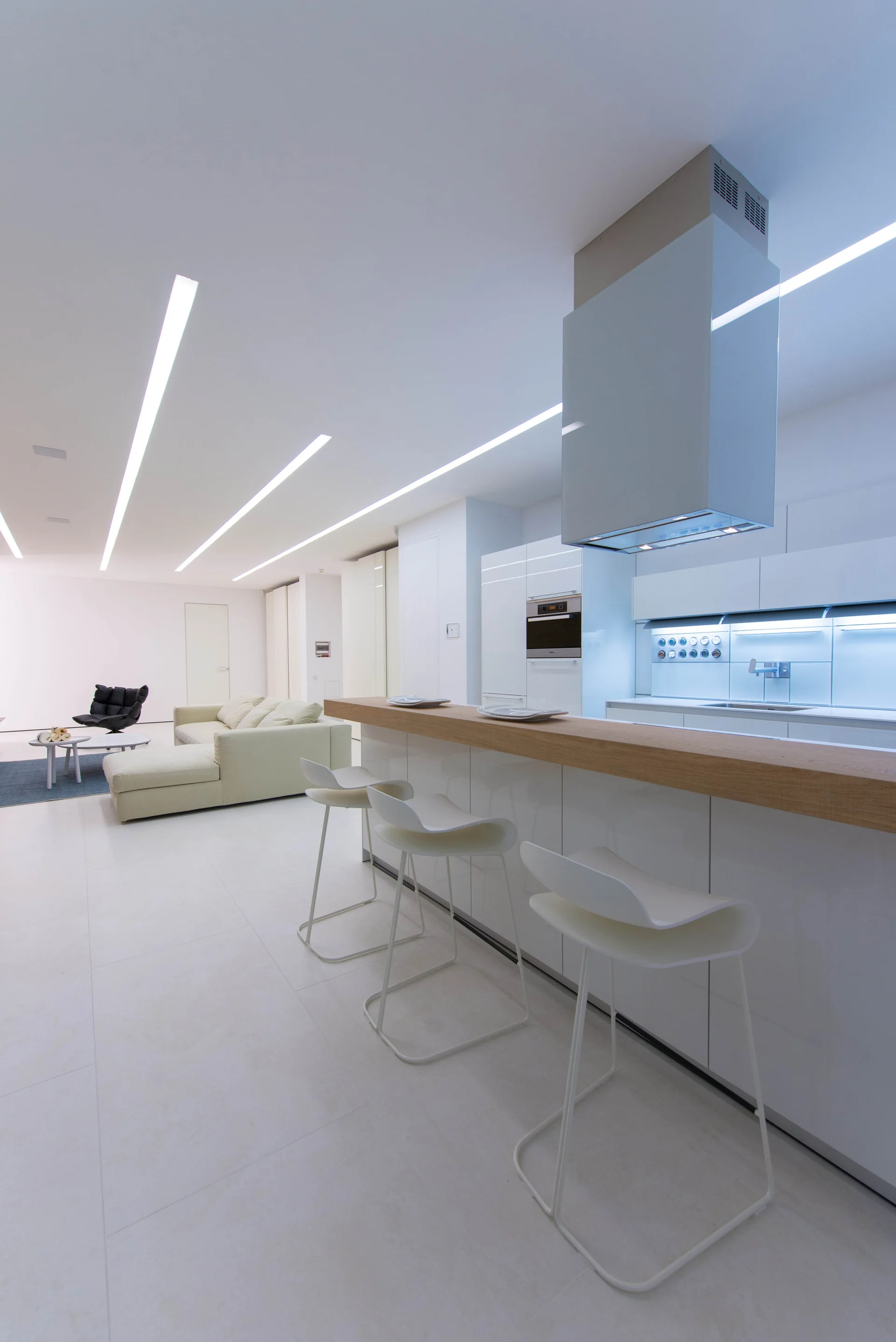 A minimalist white kitchen features a wood-topped island with three white bar stools, high-gloss cabinets, and linear ceiling lights.